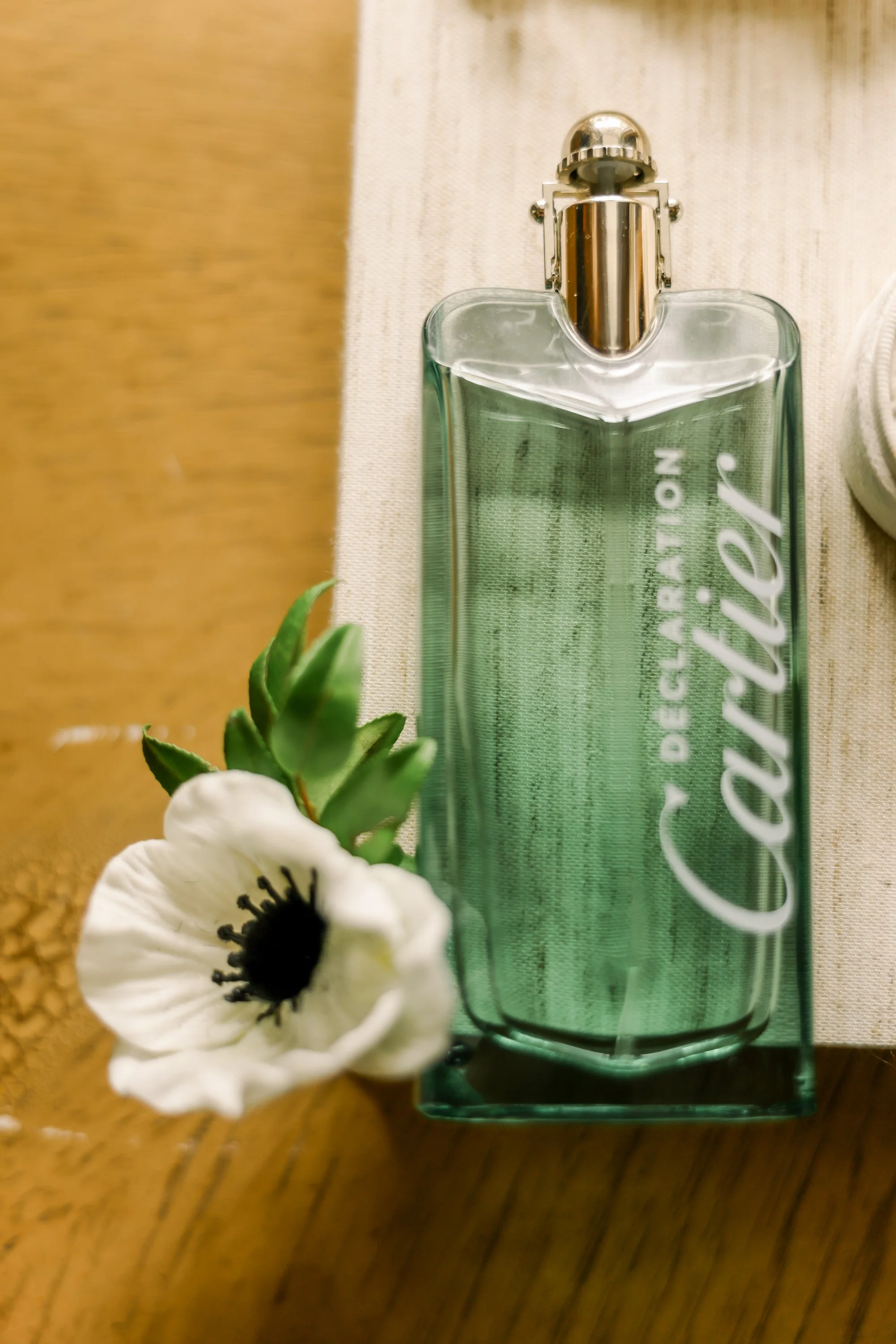 A clear rectangular perfume bottle with a silver spray nozzle, labeled 'DECLARATION' and 'Calita.' A white flower with a black center and a small cluster of green leaves are next to the bottle, on a wooden surface with a beige napkin underneath.