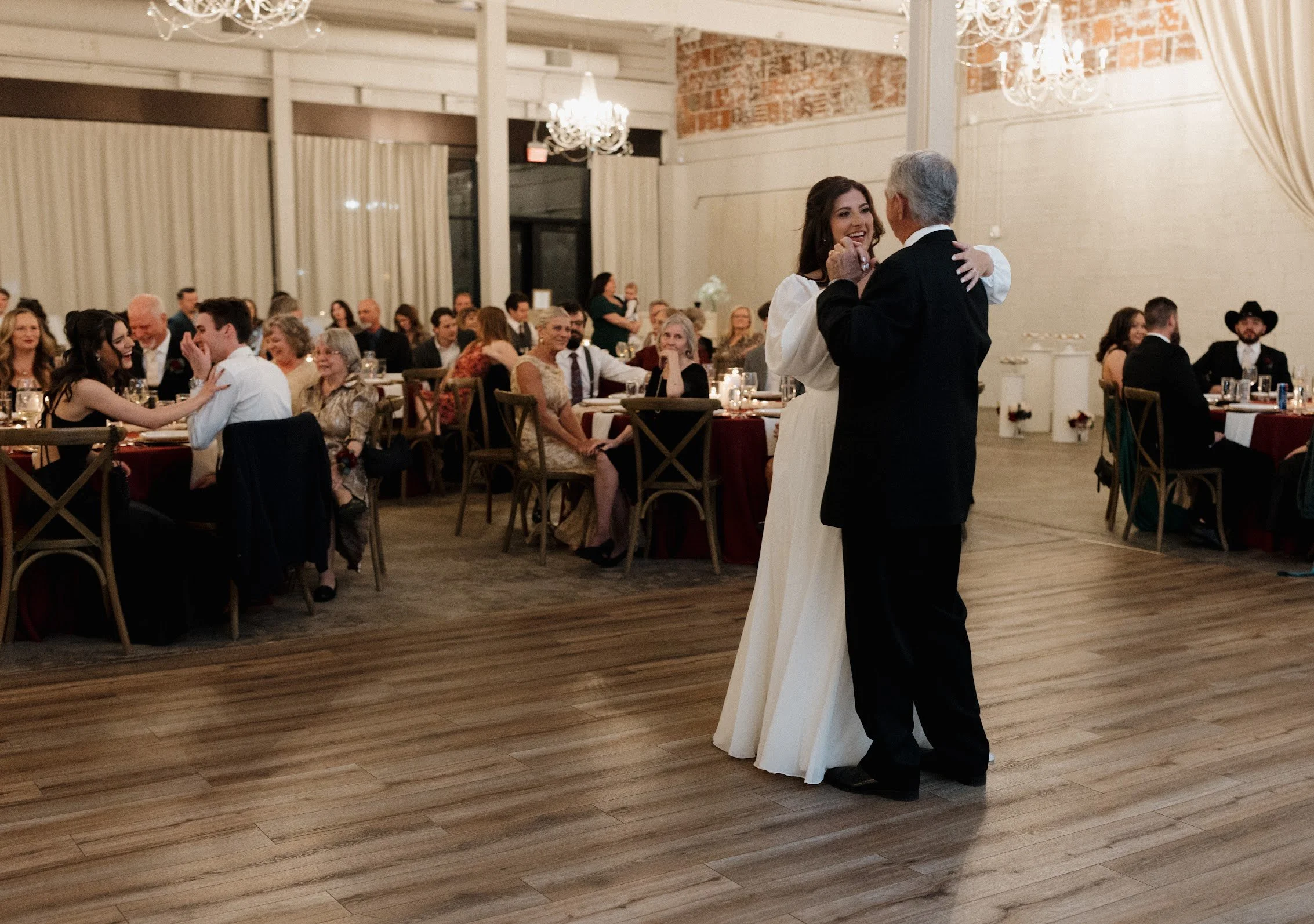 A bride and an older man are dancing in the center of a wedding reception hall filled with seated guests watching and smiling.