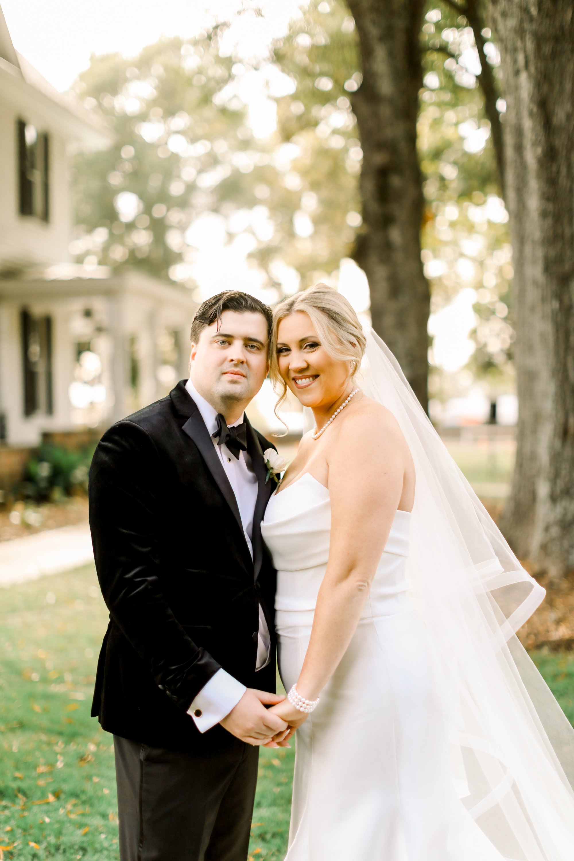 A newlywed couple holding hands outdoors on a sunny day, with trees and a house in the background. The bride is smiling and wearing a white wedding dress and pearl necklace, while the groom is in a black tuxedo with a bow tie.