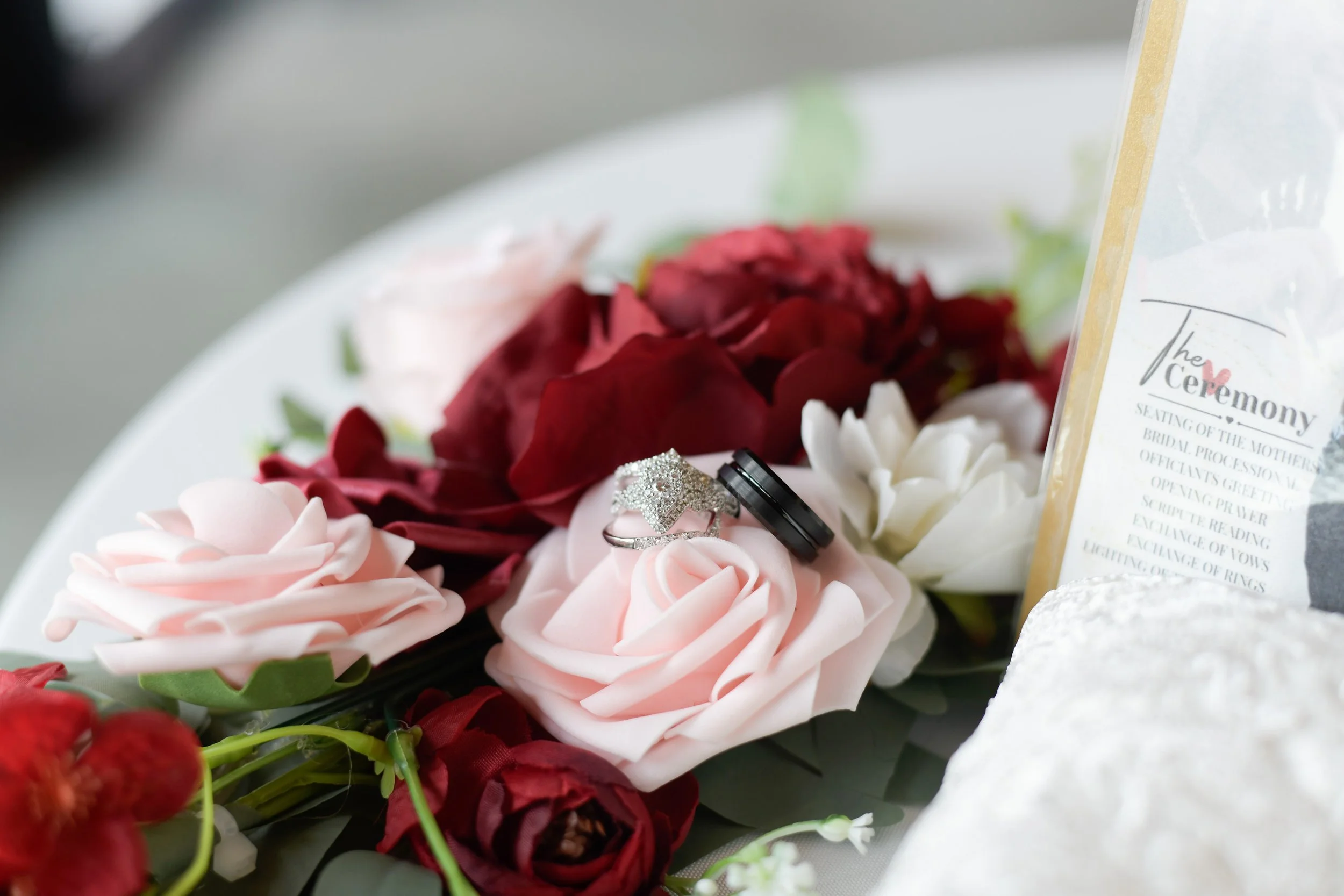 Wedding rings resting on pink and red artificial roses, with a wedding program on the right side.