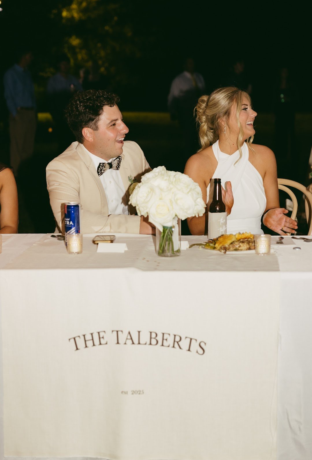 At a wedding reception, a man in a beige tuxedo with a bow tie and a woman in a white dress are sitting at a decorated table, laughing. The table has a white tablecloth, a floral centerpiece, and drinks including a can and a bottle. The tablecloth ha