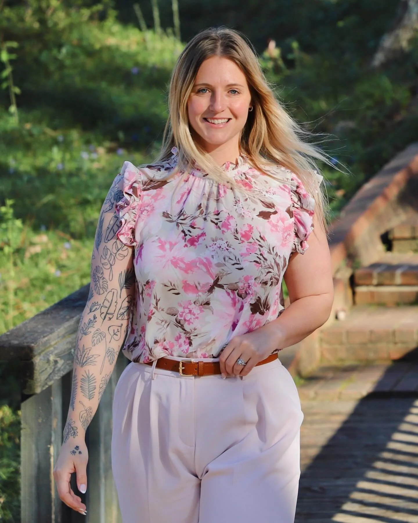 A woman with long blonde hair smiling, standing outdoors on a wooden bridge with plants in the background, wearing a floral blouse and white pants.