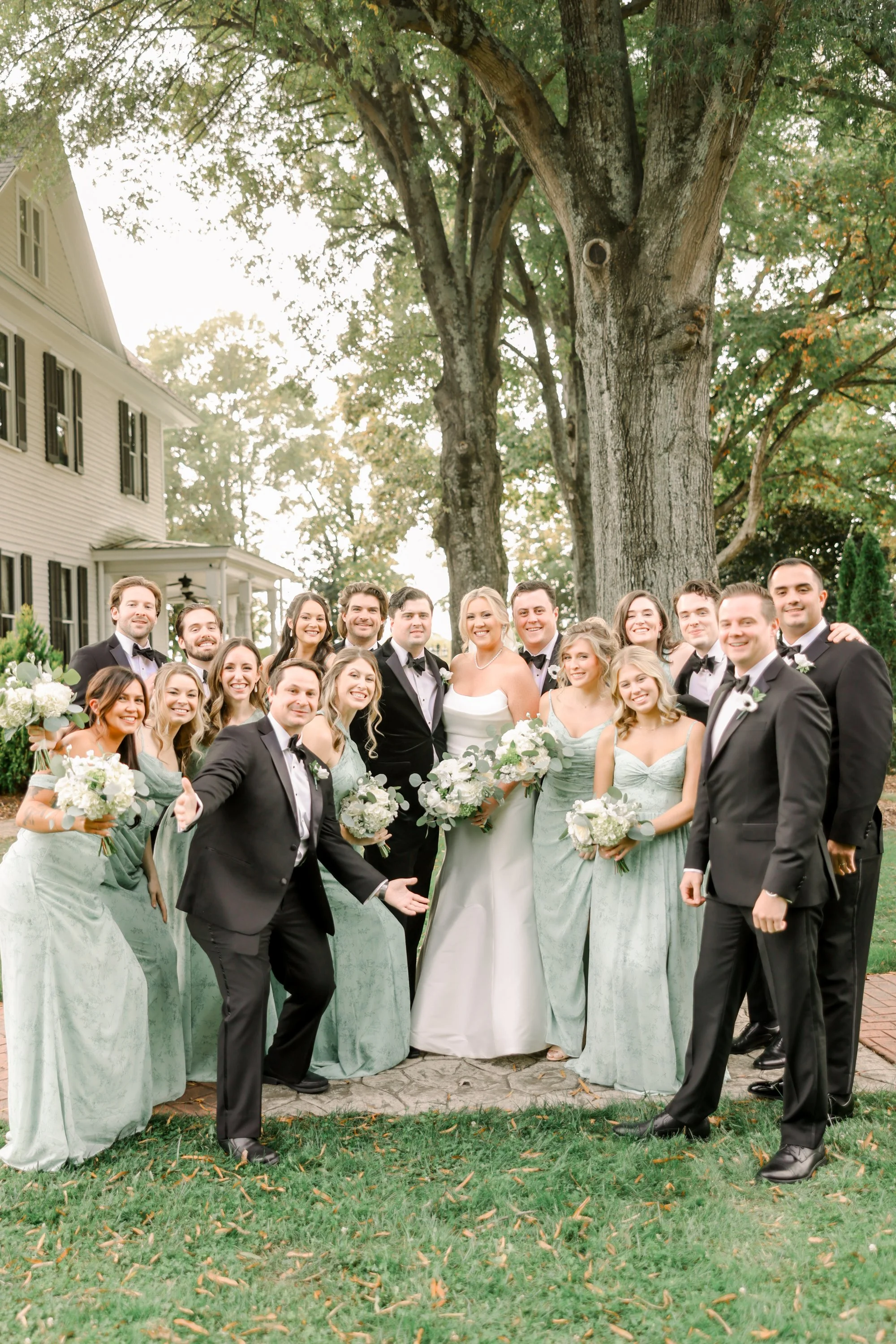 Group of people dressed in wedding attire, posing outdoors in front of a large tree and a house.