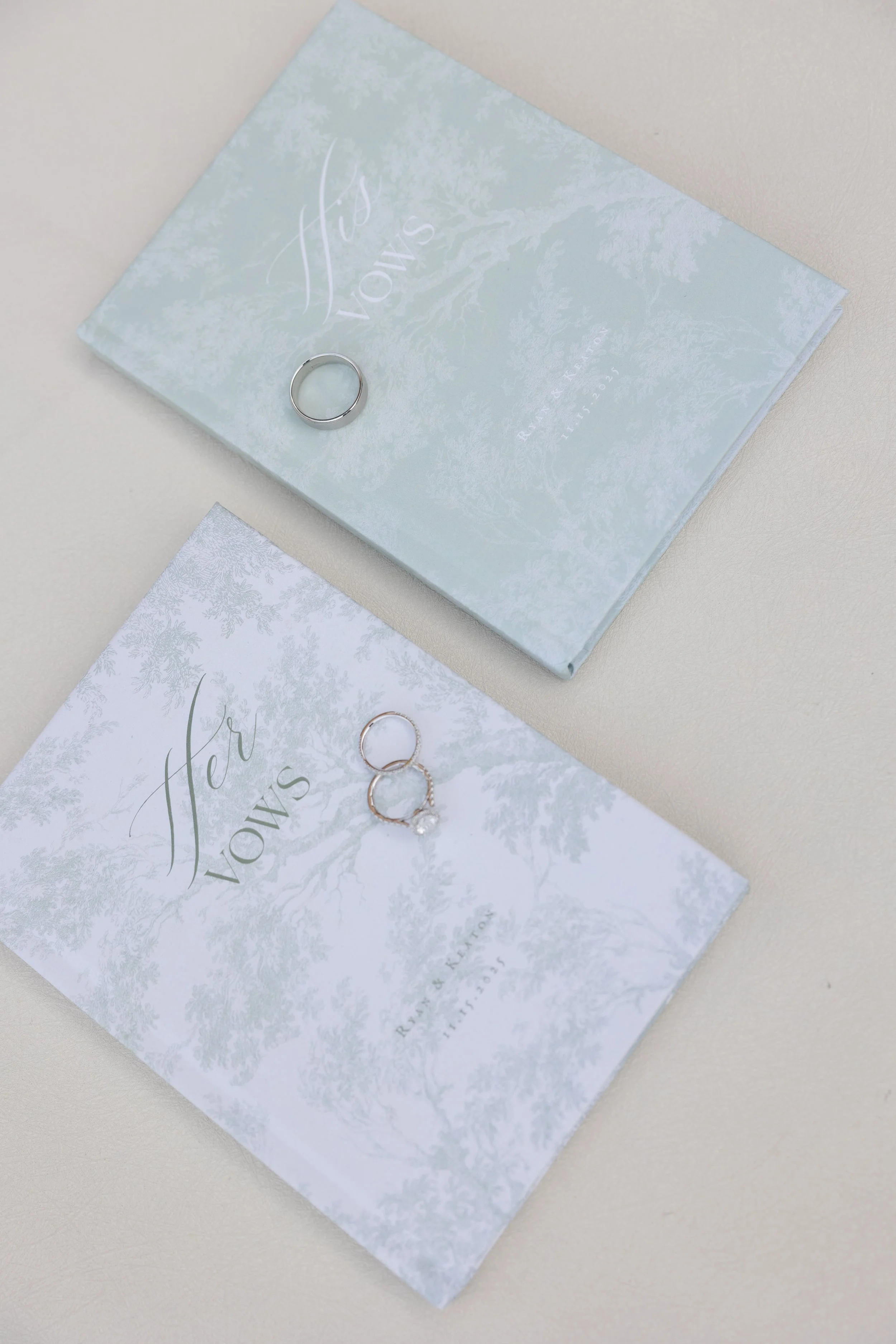 A wedding ring and two engagement rings on decorative paper with text that says "Her Vows" and a matching blue box, set on a light-colored surface.