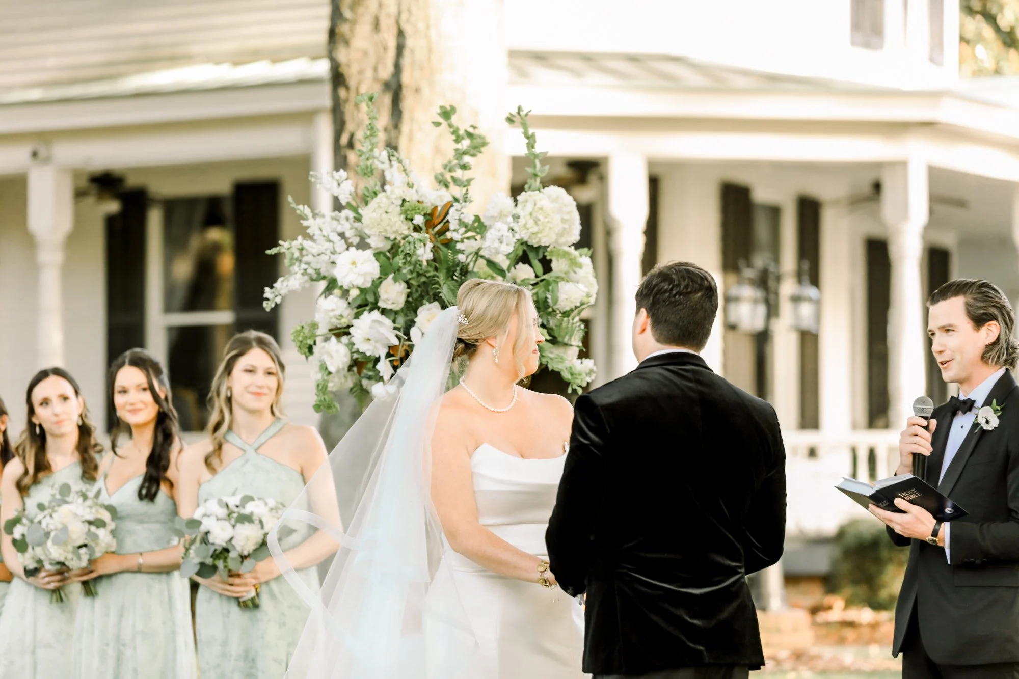 A couple gets married outdoors, with a bride in a white gown and veil, a groom in a black tuxedo, and an officiant reading vows. Bridesmaids in matching light green dresses hold bouquets, and there is a large floral arrangement with white flowers beh