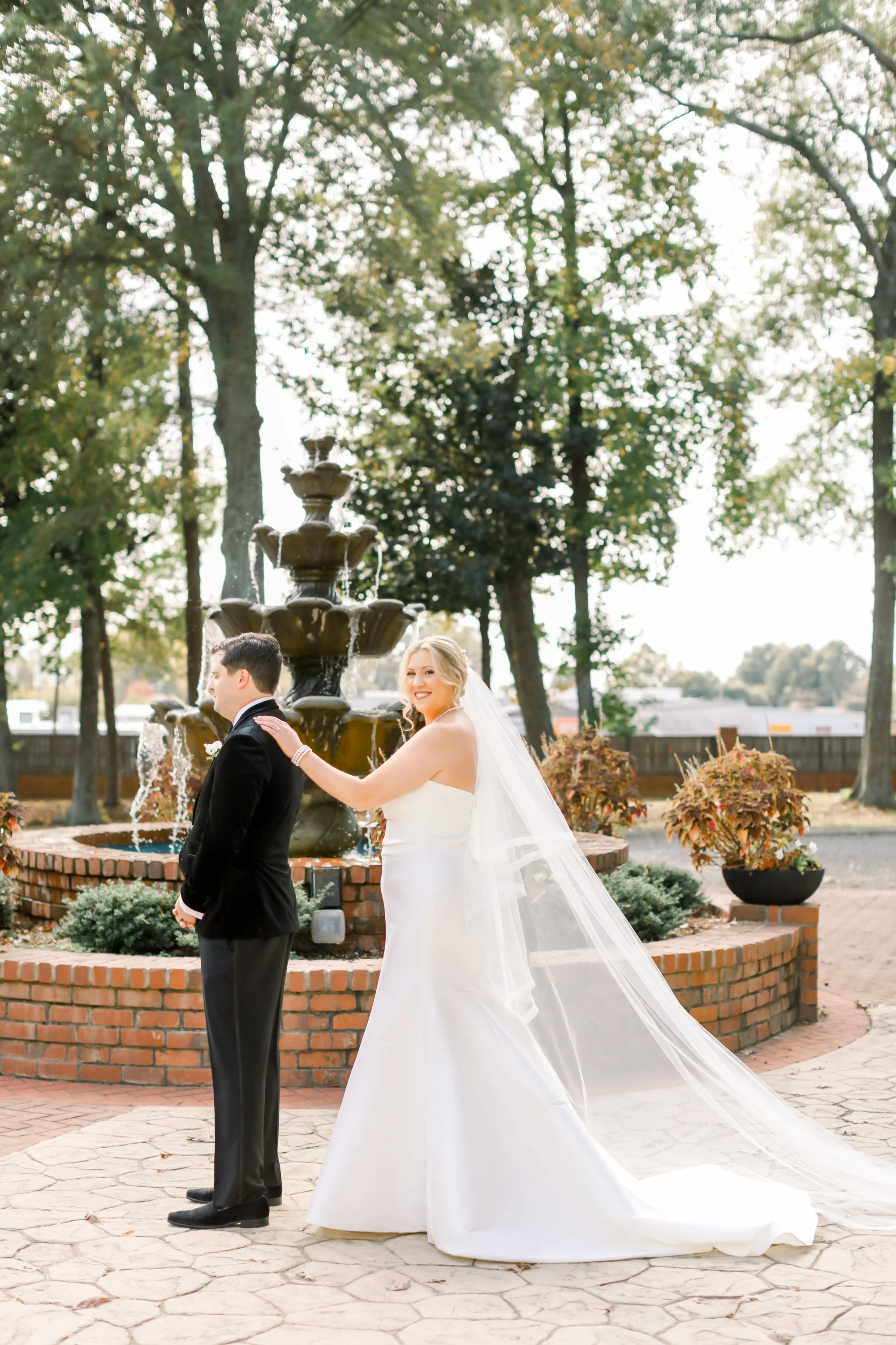 Bride in white wedding dress touching groom in black tuxedo in front of outdoor fountain with trees and plants.