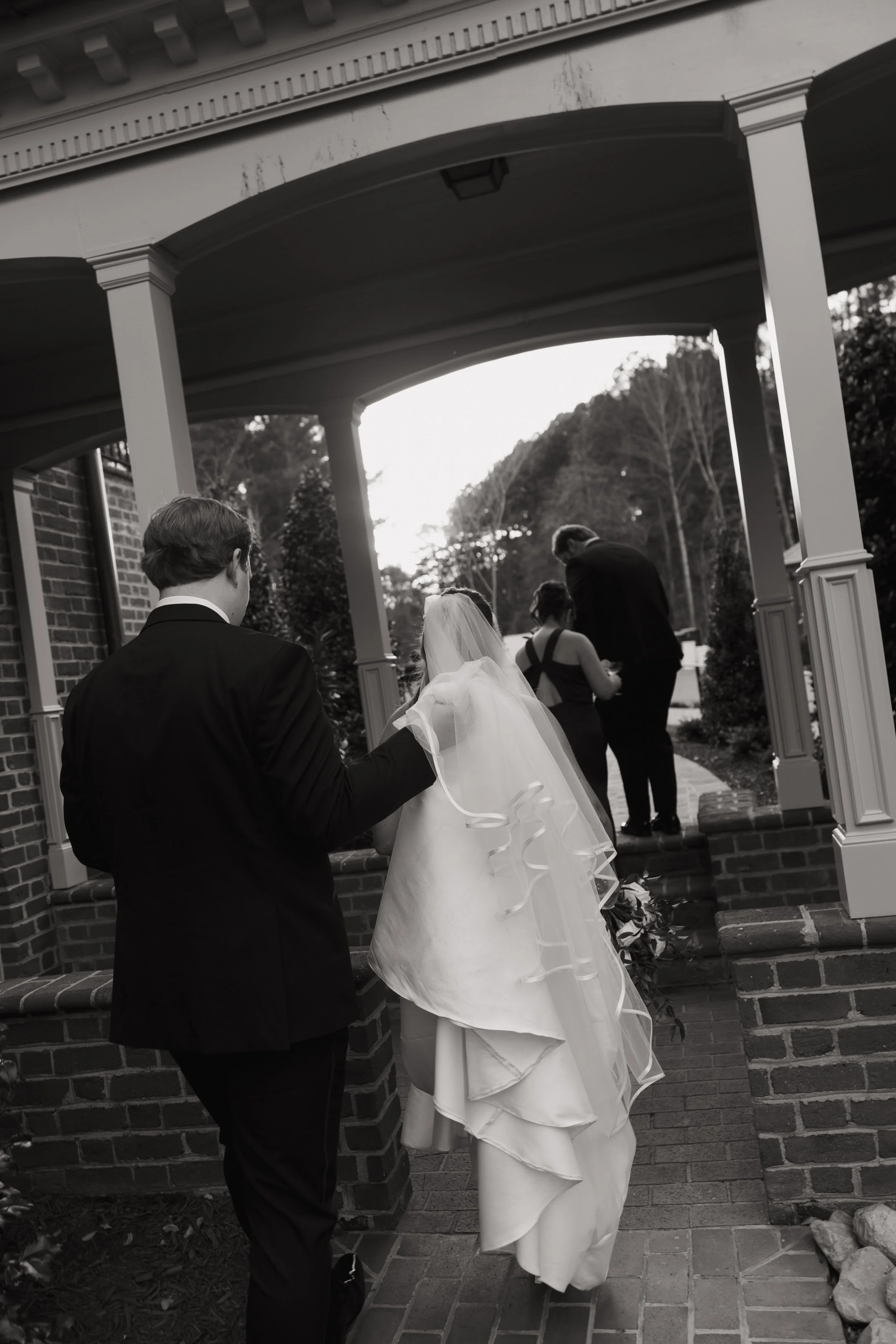 A black-and-white photo of a bride and groom walking together, with the groom holding the bride's veil, outside a house with a porch and brick steps.
