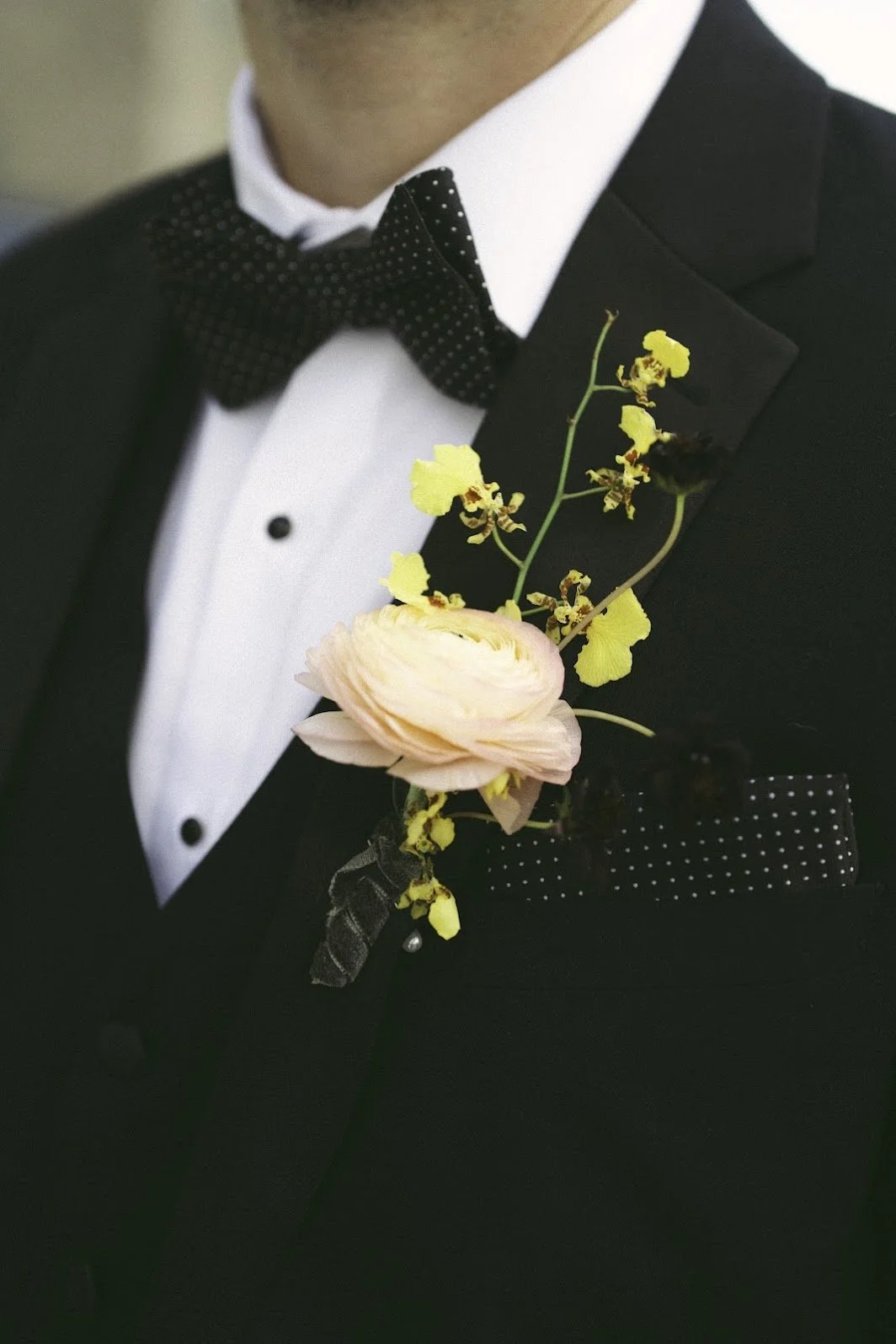 A man in a tuxedo with a floral boutonniere on his lapel, featuring a pink ranunculus and yellow orchids.