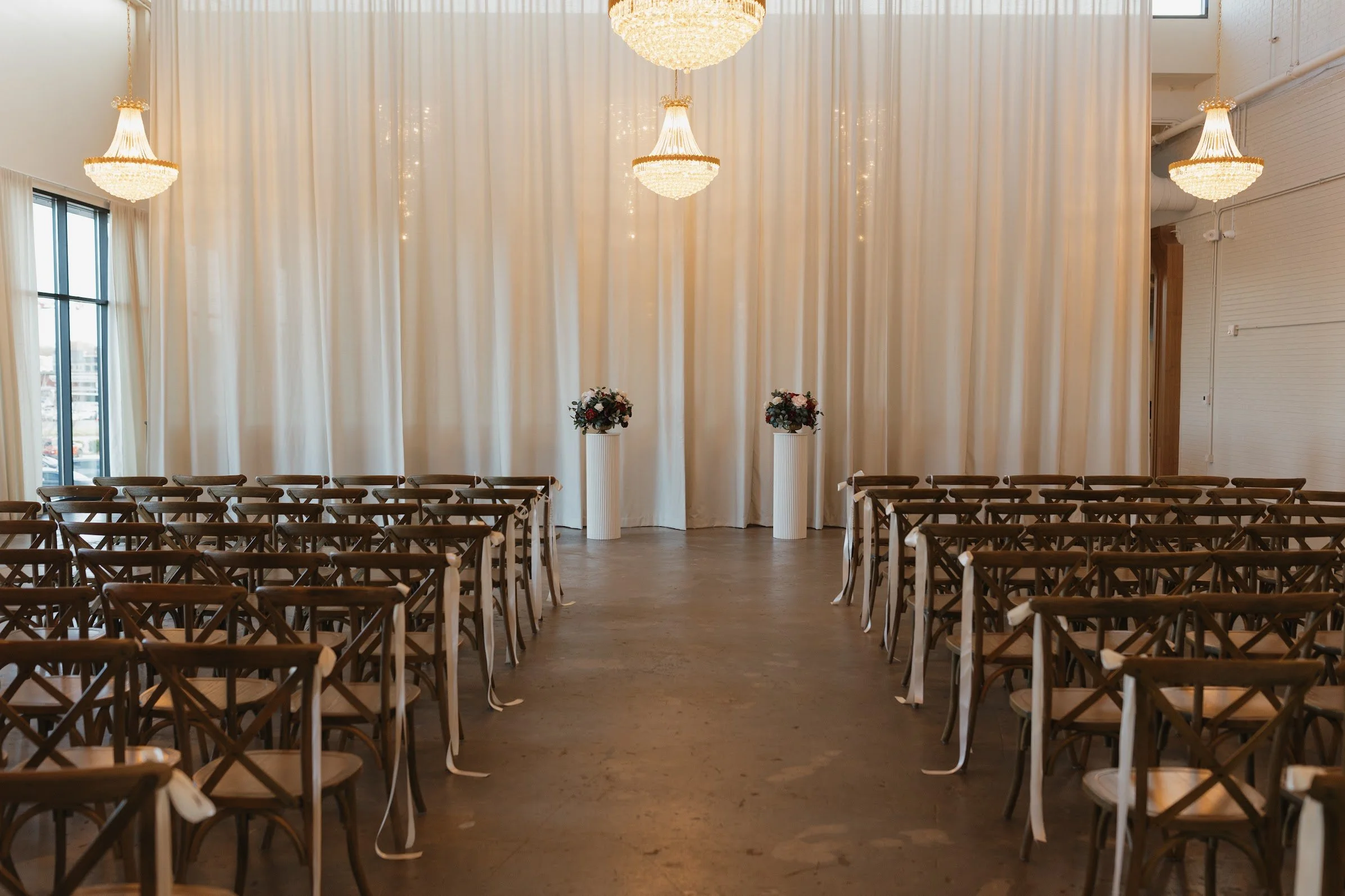 Empty wedding ceremony space with chairs arranged on both sides, two flower arrangements on white pedestals, a cream-colored curtain backdrop, and chandeliers hanging from the ceiling.