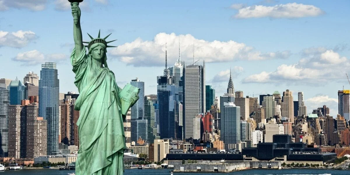 Estatua de la libertad en la ciudad de Nueva York con edificios del skyline al fondo y cielo con nubes.