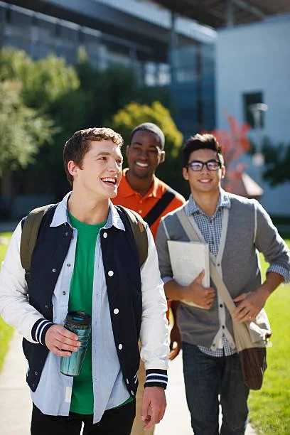 Tres estudiantes universitarios caminando y sonriendo en un campus, uno lleva una mochila y un vaso con bebida, otro sostiene libros y todos parecen felices.