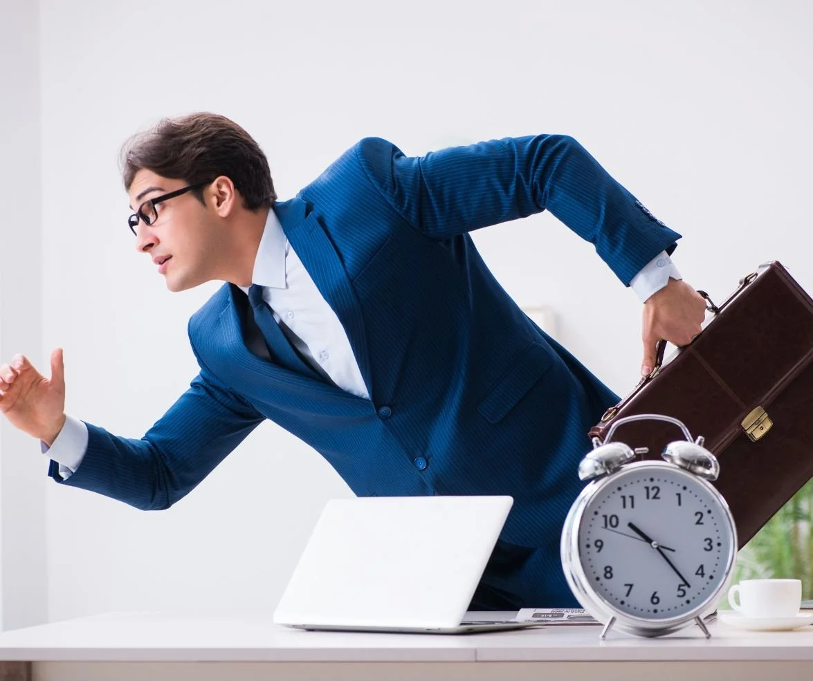 Hombre de negocios vestido con traje azul y gafas, enojado, atrapado en un reloj de alarma gigante, en una oficina moderna.