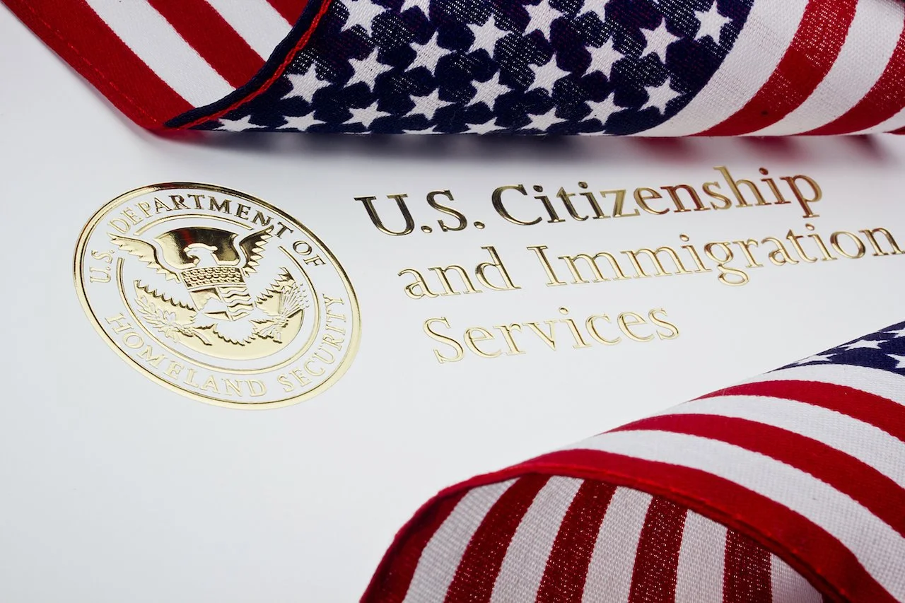 Documento con texto dorado que dice 'U.S. Citizenship and Immigration Services' y un sello dorado del Departamento de Seguridad Nacional de EE. UU., rodeado de cinta con estrellas y rayas características de la bandera de Estados Unidos.