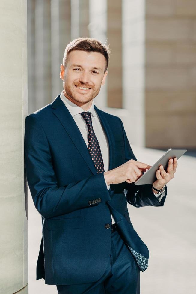 Hombre de negocios sonriendo, usando traje azul y corbata, apoyado en una pared y usando una tablet en un ambiente moderno.
