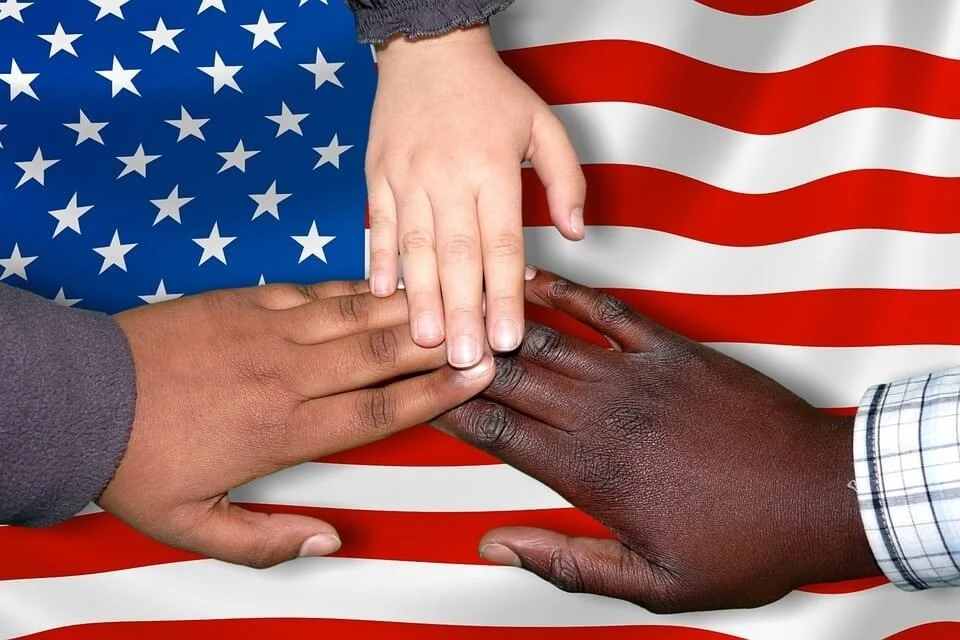Tres manos de diferentes tonos de piel tocándose sobre la bandera de Estados Unidos.