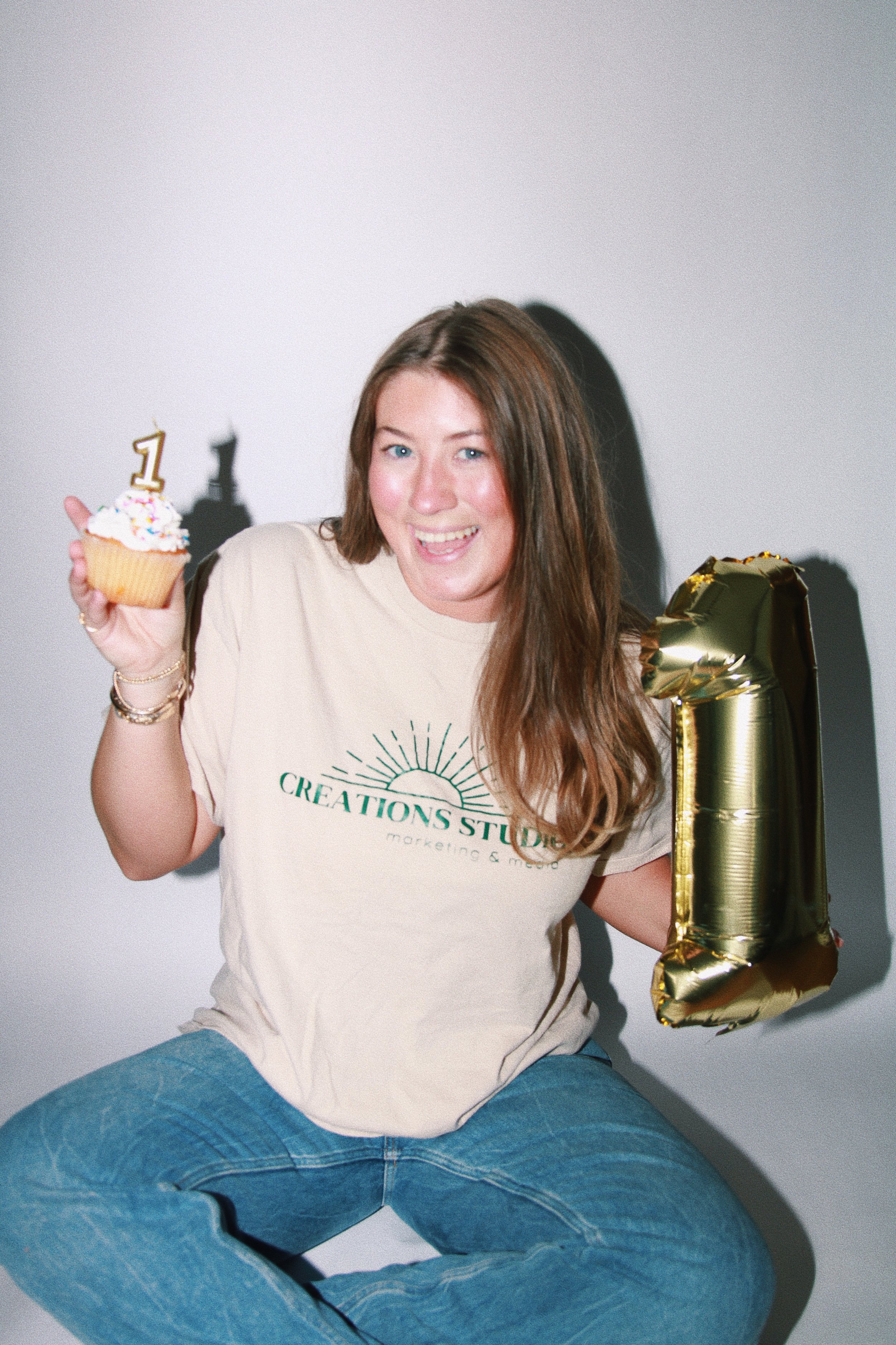 Woman sitting, holding a cupcake with a number 1 candle, and a large gold number 1 balloon, celebrating her first birthday.