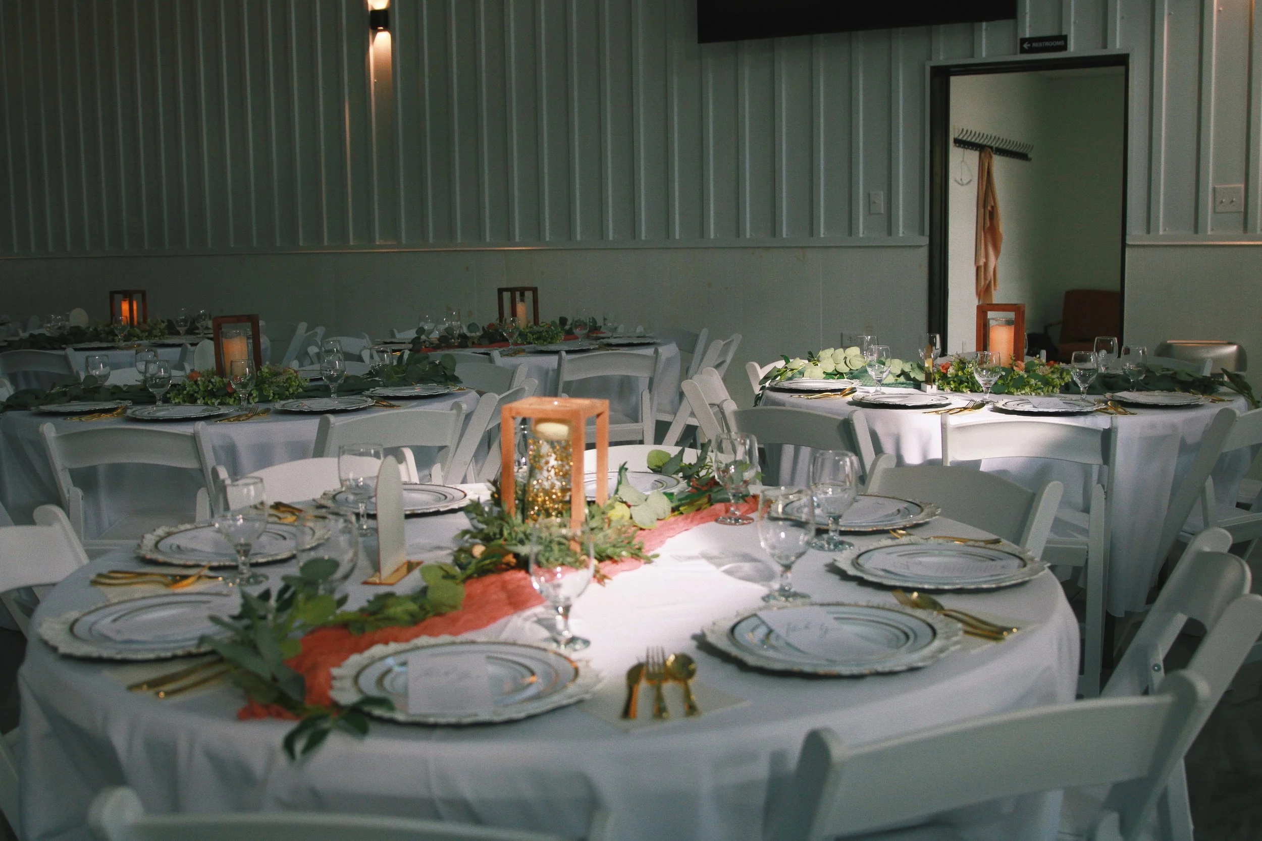 Empty banquet tables set for a wedding or formal event with white tablecloths, gold flatware, glassware, and floral centerpieces, in a decorated hall.