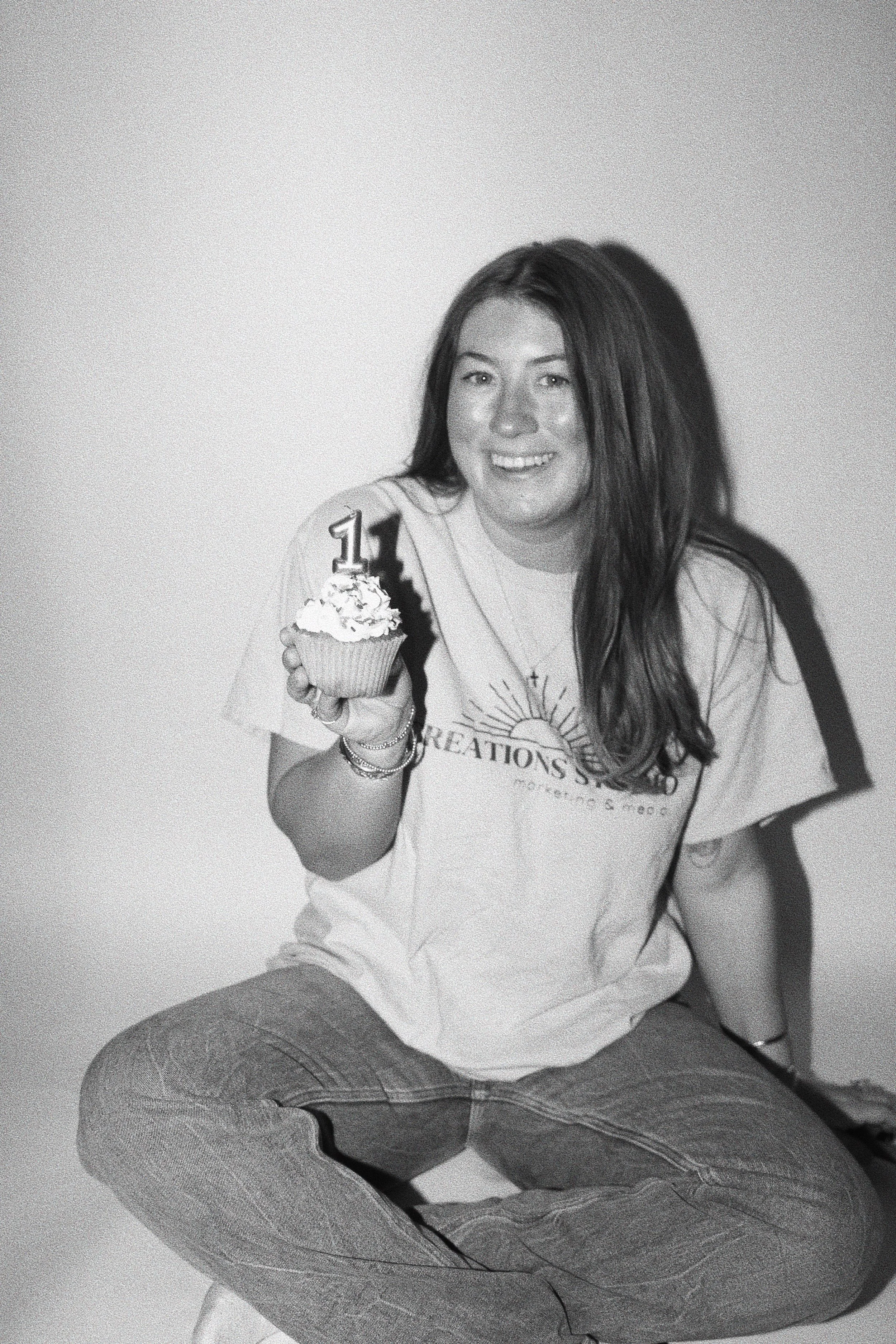 A woman with long dark hair sitting cross-legged and holding a cupcake with a birthday candle that reads '11', smiling at the camera, wearing a t-shirt.