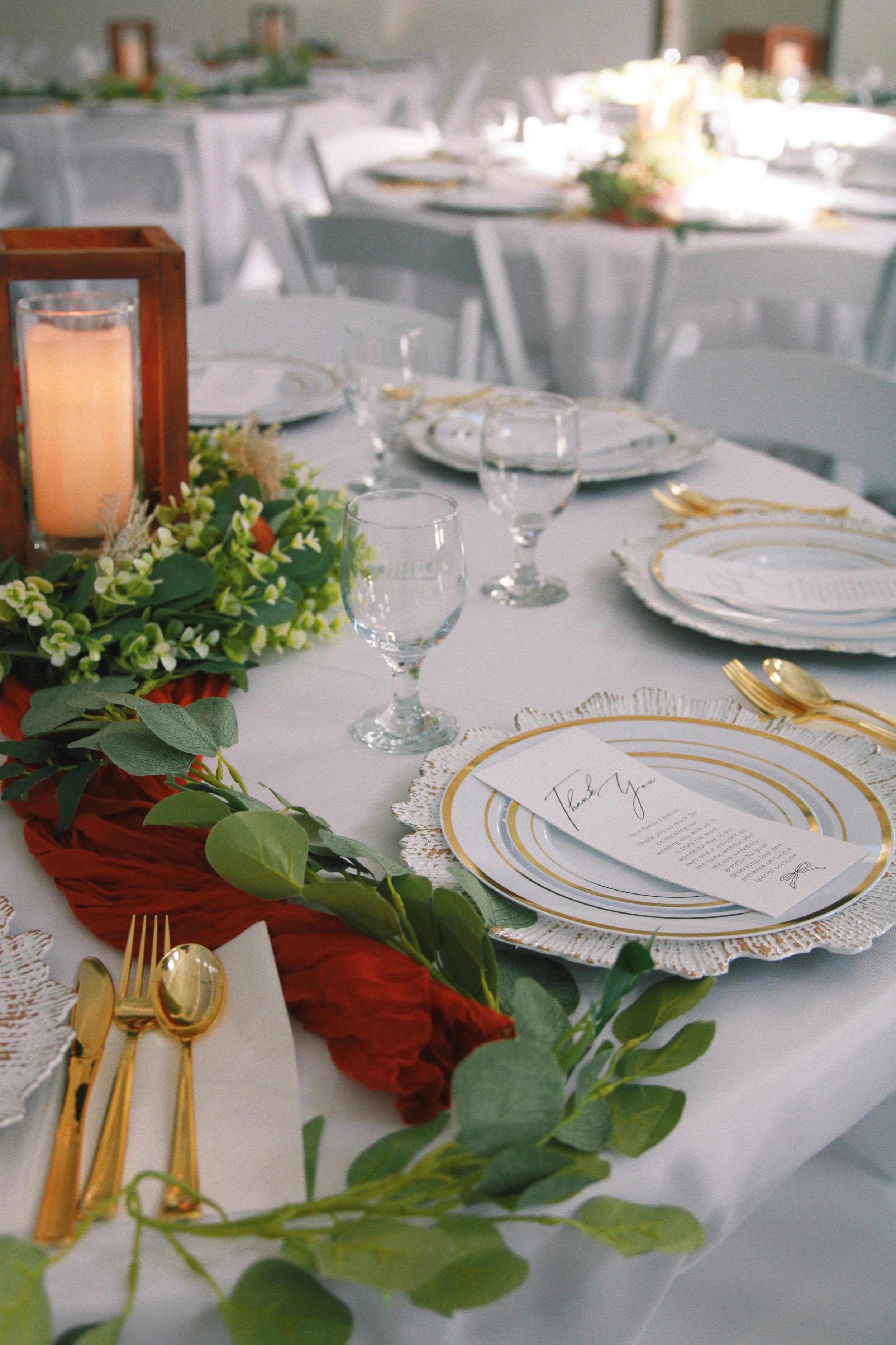 Elegant banquet table setup with white tablecloth, gold flatware, glassware, decorative greenery, and a white 'Thank You' note on a plate, in a well-lit event space.