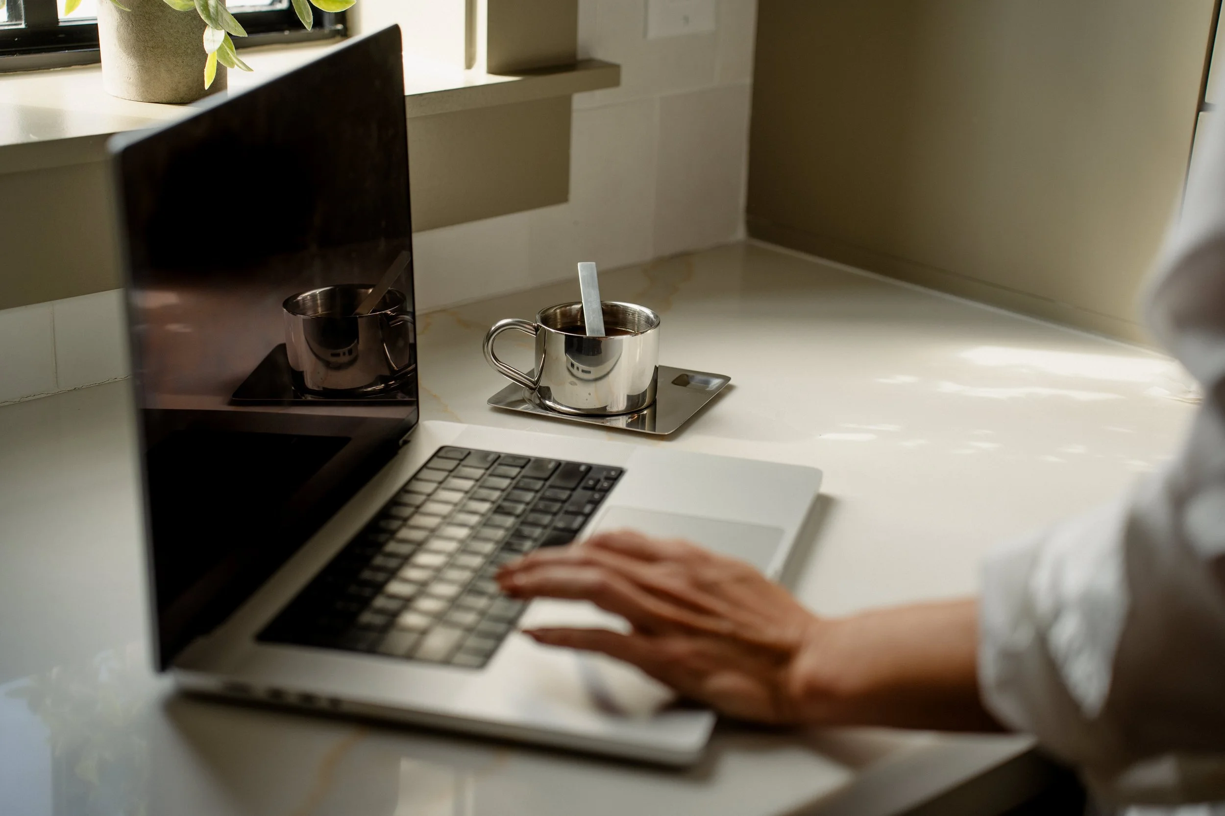 Person working on a laptop at a white countertop next to a window with sunlight and a potted plant, with a metal cup containing a stir stick nearby.