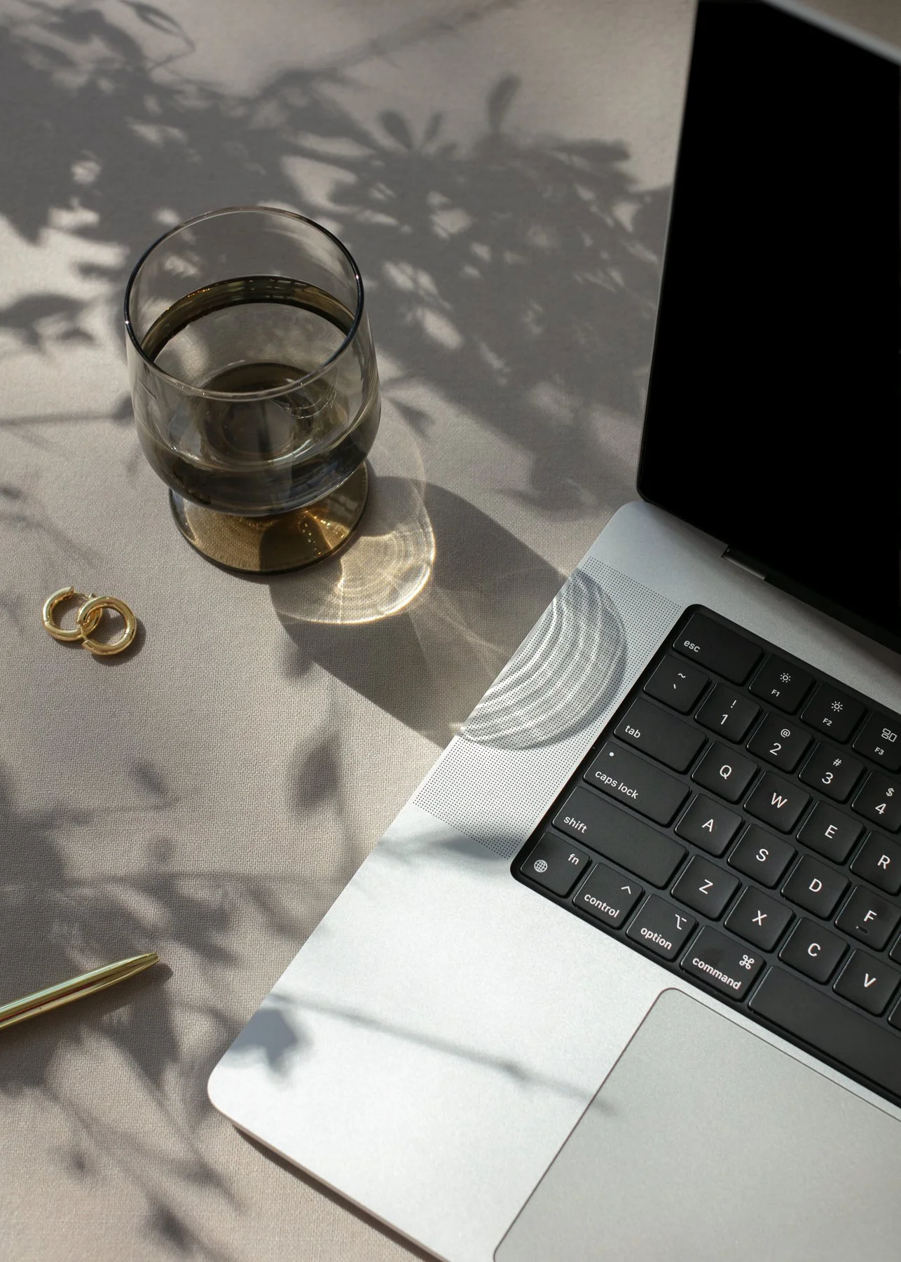 A close-up of a silver laptop computer, a glass of water with a golden base, a pair of gold earrings, and a gold pen on a white tablecloth with shadows of leaves.
