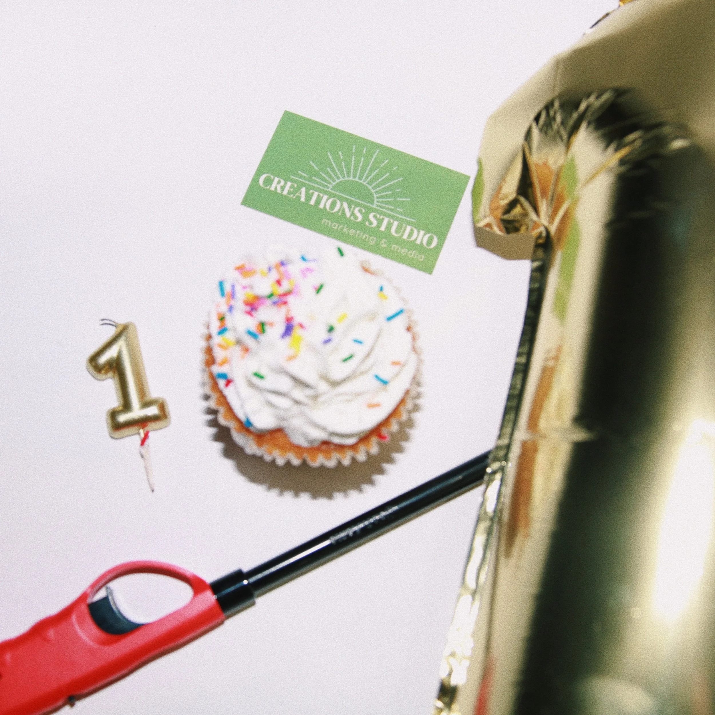 A cupcake with white frosting and colorful sprinkles on a white surface, next to a gold-colored number one candle, a green business card from 'Creations Studio Marketing & Media,' and a small section of a wrapped gift with shiny gold paper.