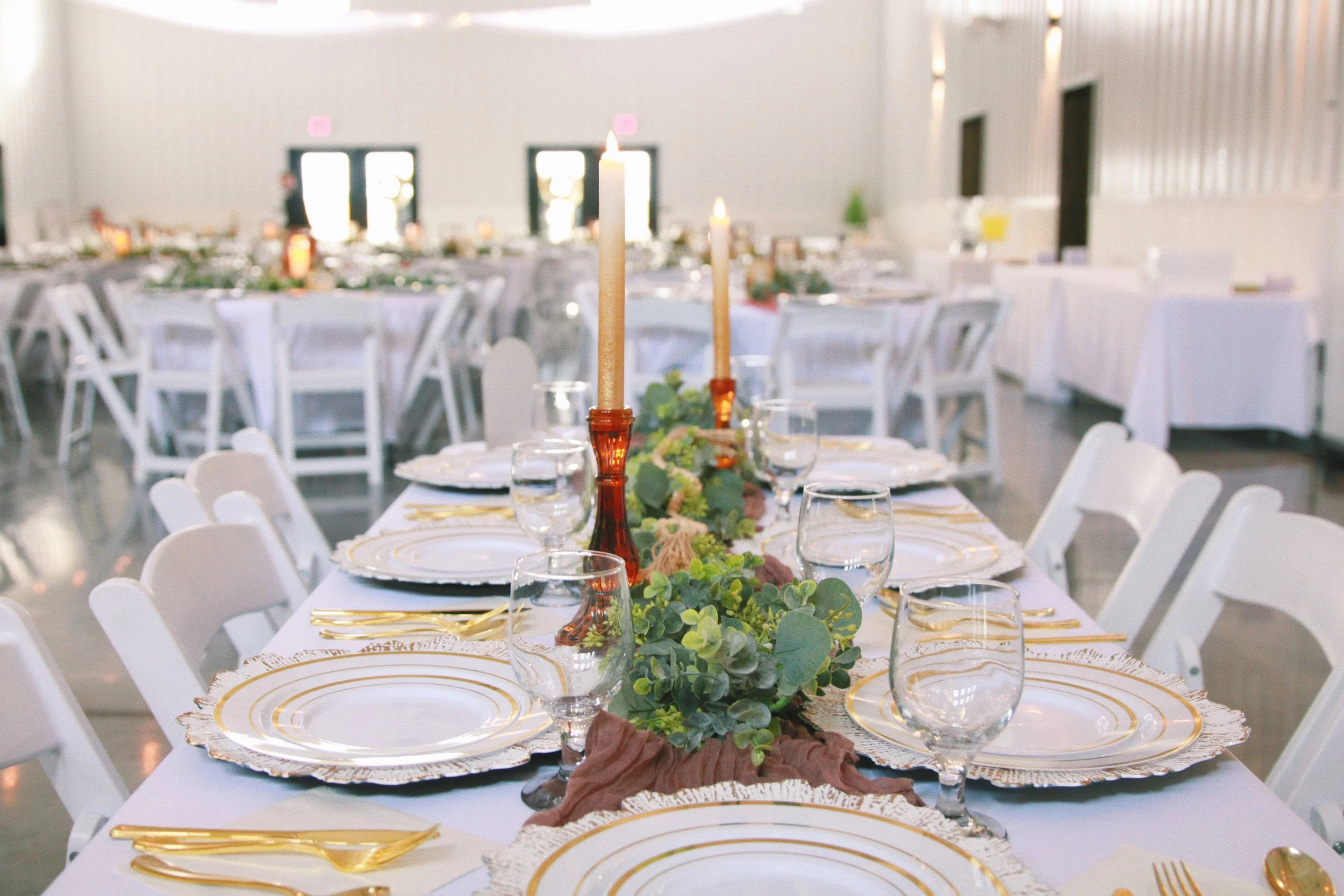 Long banquet table set for a formal event with white tablecloths, gold-rimmed plates, gold cutlery, wine glasses, candles, and greenery centerpieces in a bright, spacious venue