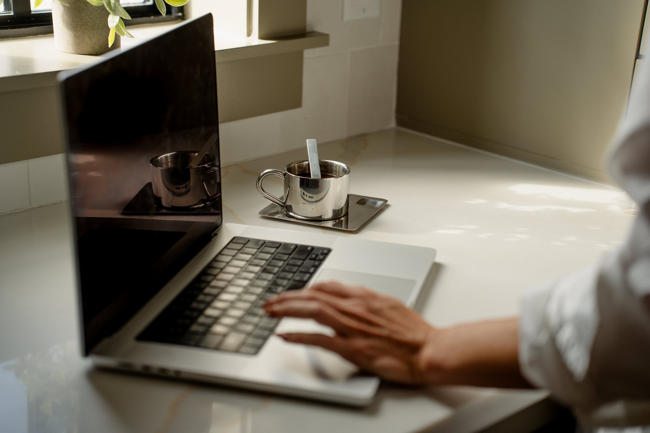 A person typing on a laptop computer on a white countertop, with a metal cup containing a stir stick and a laptop showing the cup's reflection nearby, in a bright room with sunlight filtering through a window.