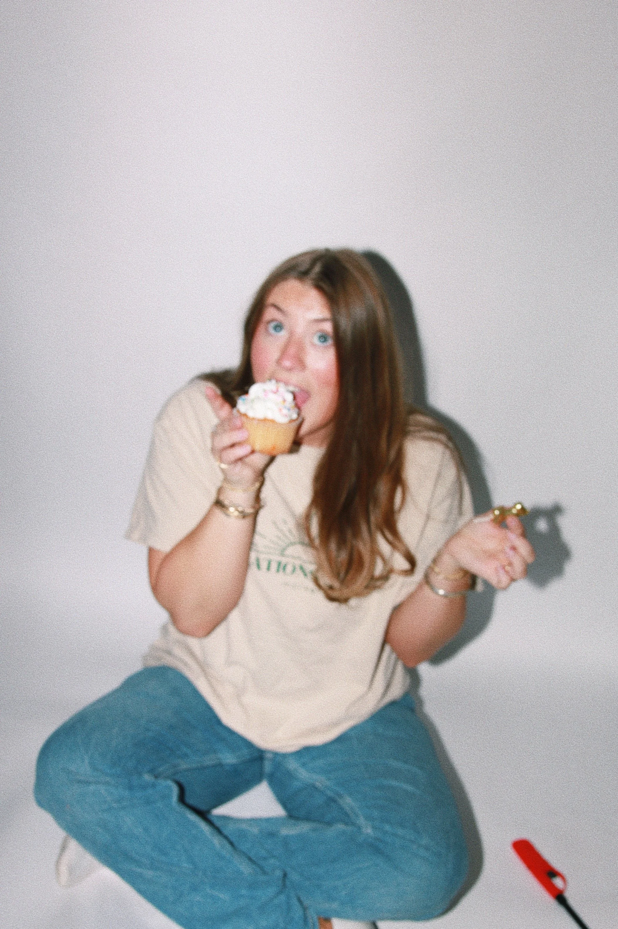 A young woman with long brown hair and blue eyes squats while holding a cupcake with white frosting and sprinkles, and a matchstick in her other hand, against a plain white background.
