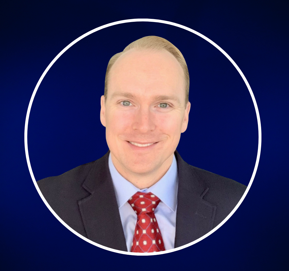 Professional headshot of a man with light skin, blonde hair, dressed in a dark suit, light blue shirt, and red patterned tie, smiling at the camera against a plain grey background.