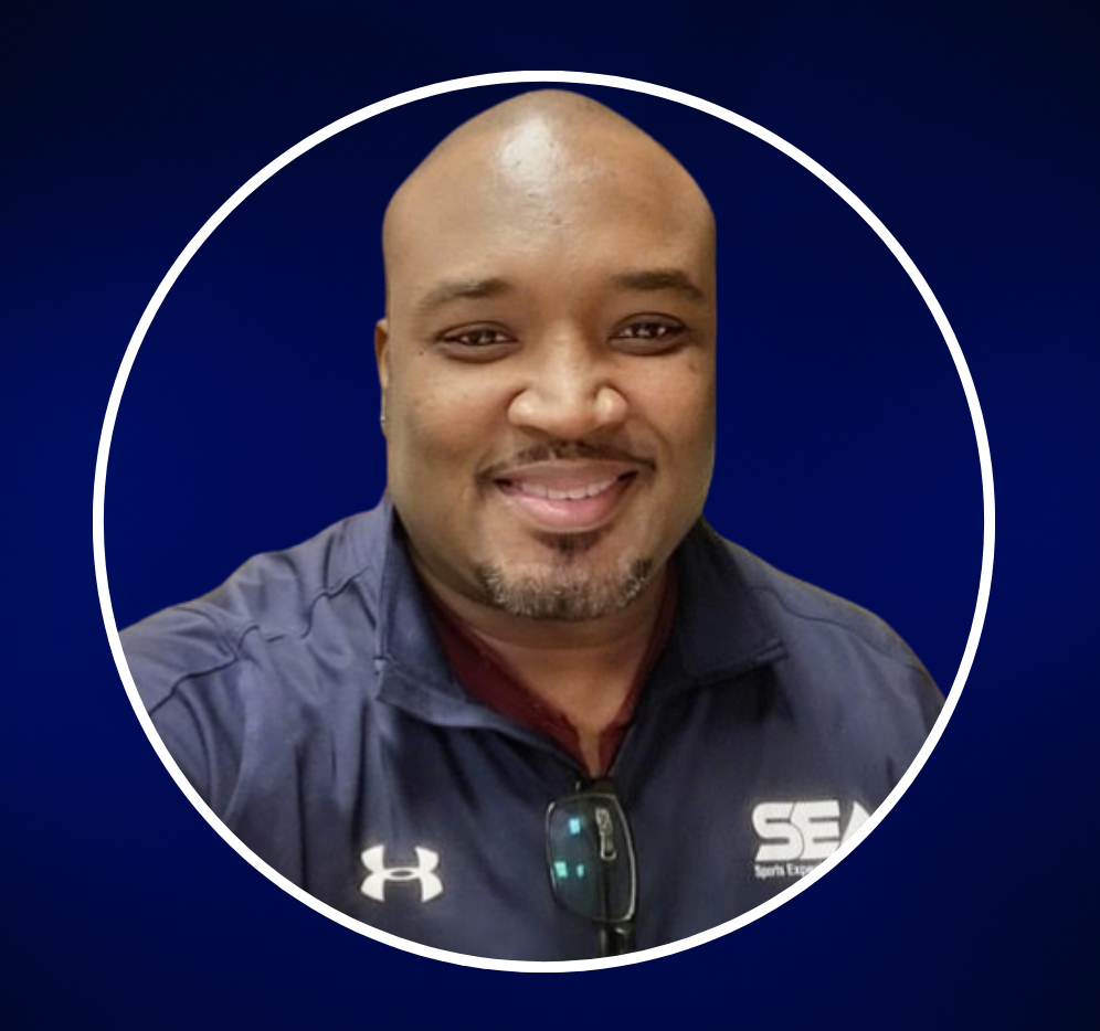 A smiling man taking a selfie against a plain dark wall, wearing a navy blue sports jacket with 'SEA' logo and sunglasses hanging from his collar.