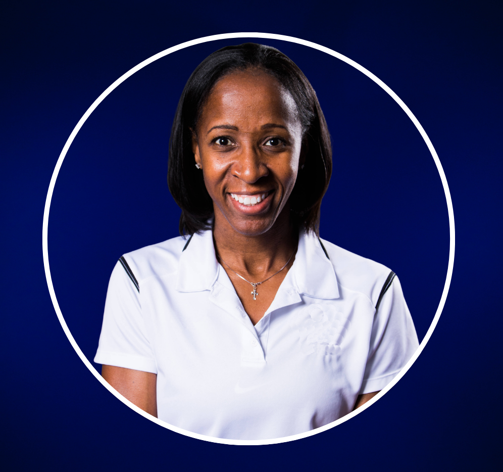 A woman with dark hair smiling, wearing a white collared shirt and a cross necklace against a gray background.