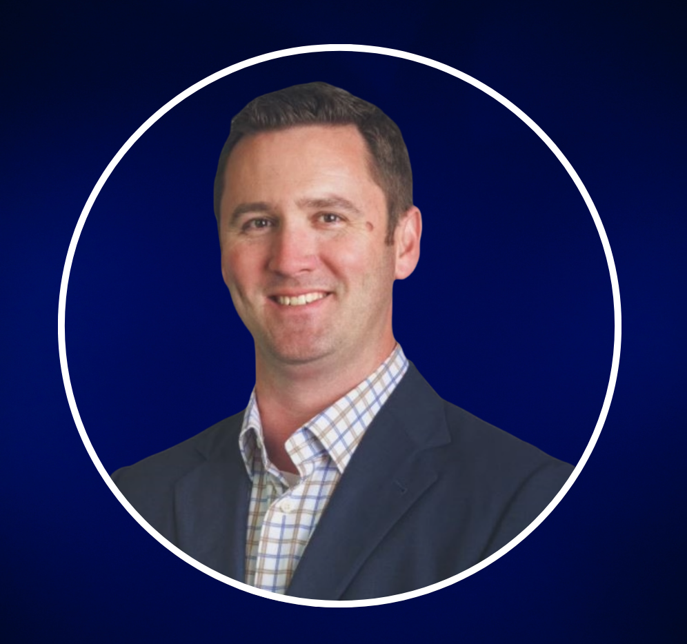 Headshot of a smiling man in a suit with a checkered shirt against a plain background.