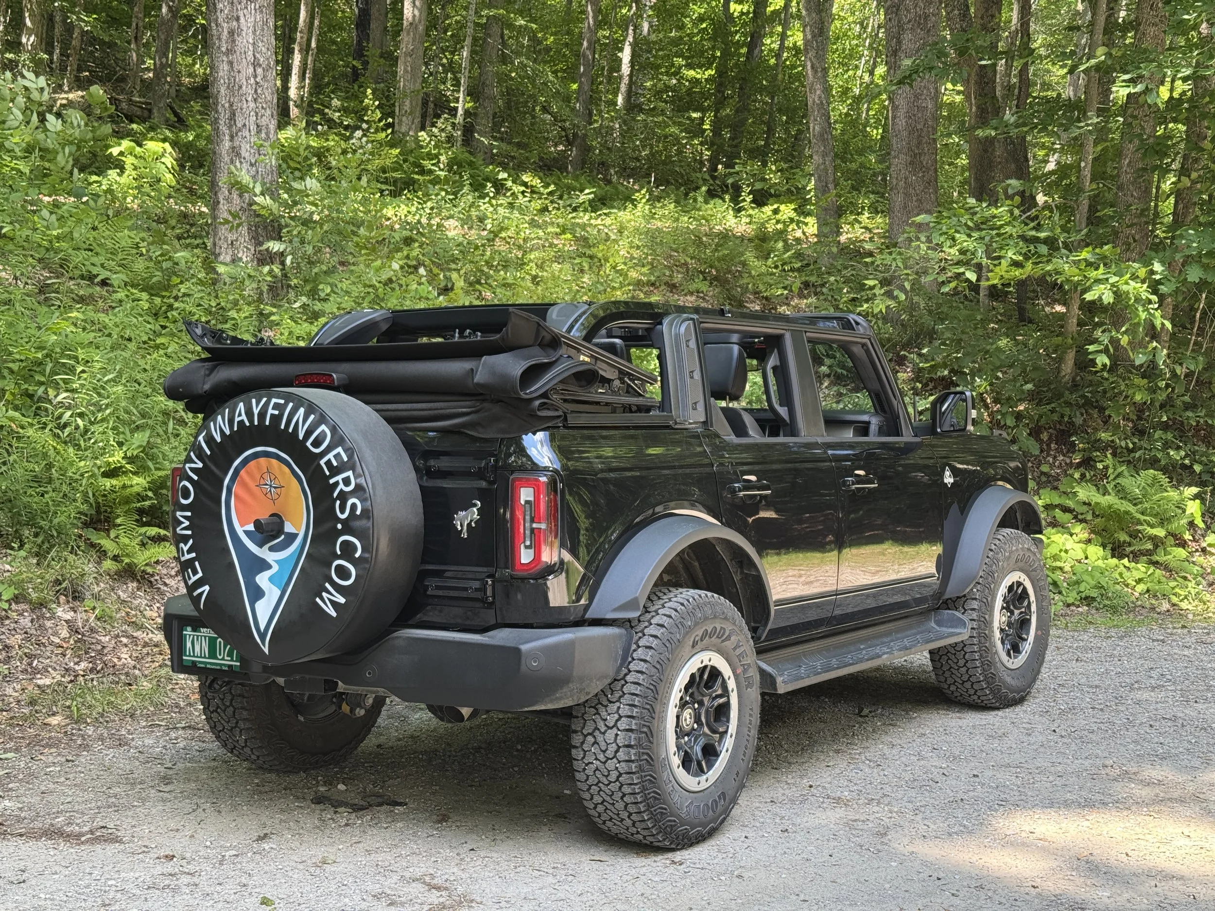 A black Bronco with tire cover reading 'Vermont Wayfinders' parked on a gravel trail in a lush green forest.