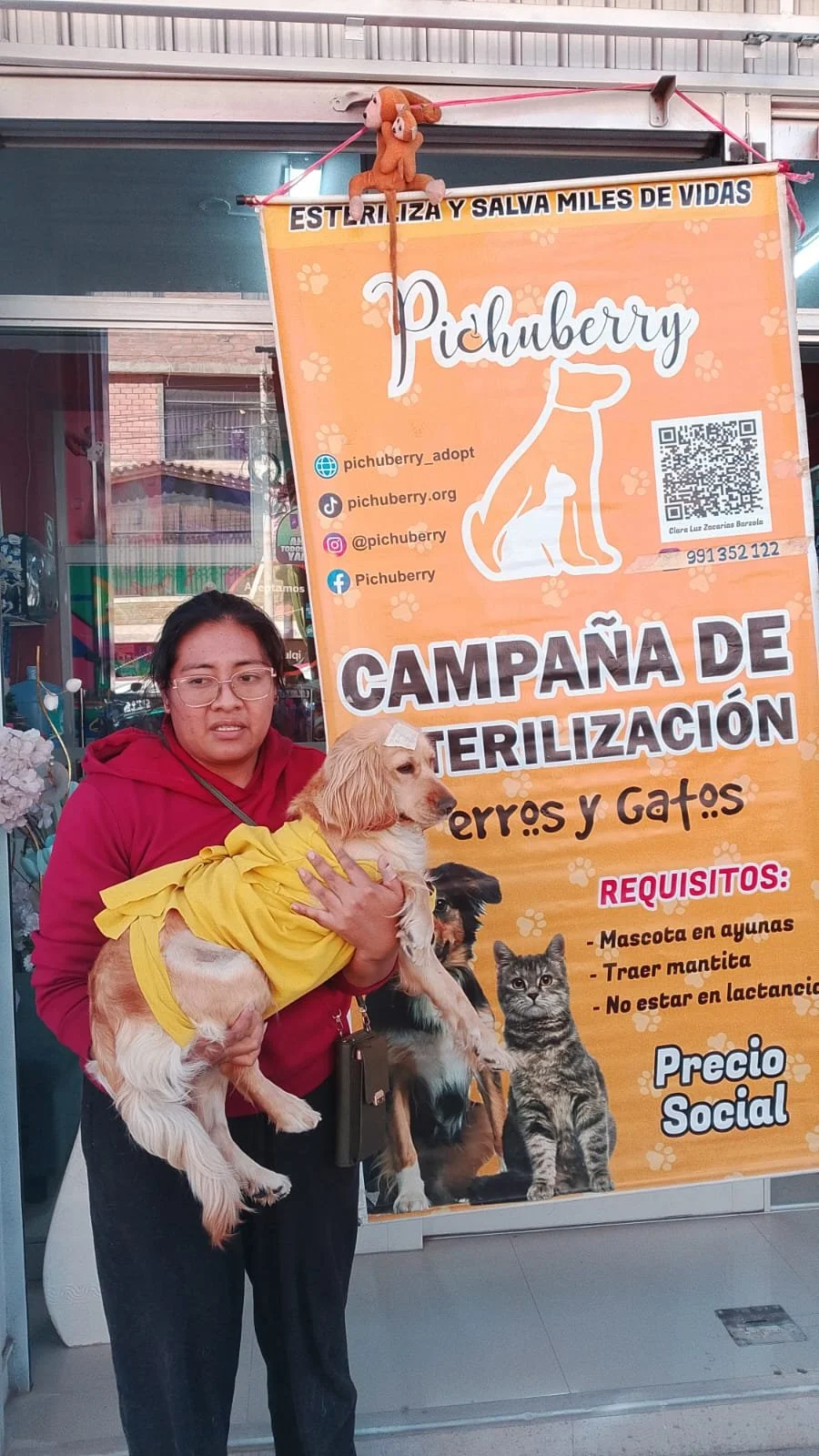 A woman in a red hoodie and glasses holding a small dog and a larger dog in front of an orange banner advertising pet sterilization campaign for dogs and cats.
