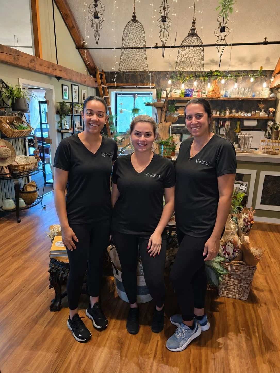Three women wearing black shirts with a logo, standing in a cozy, well-decorated shop or cafe with plants, shelves, and hanging light fixtures.