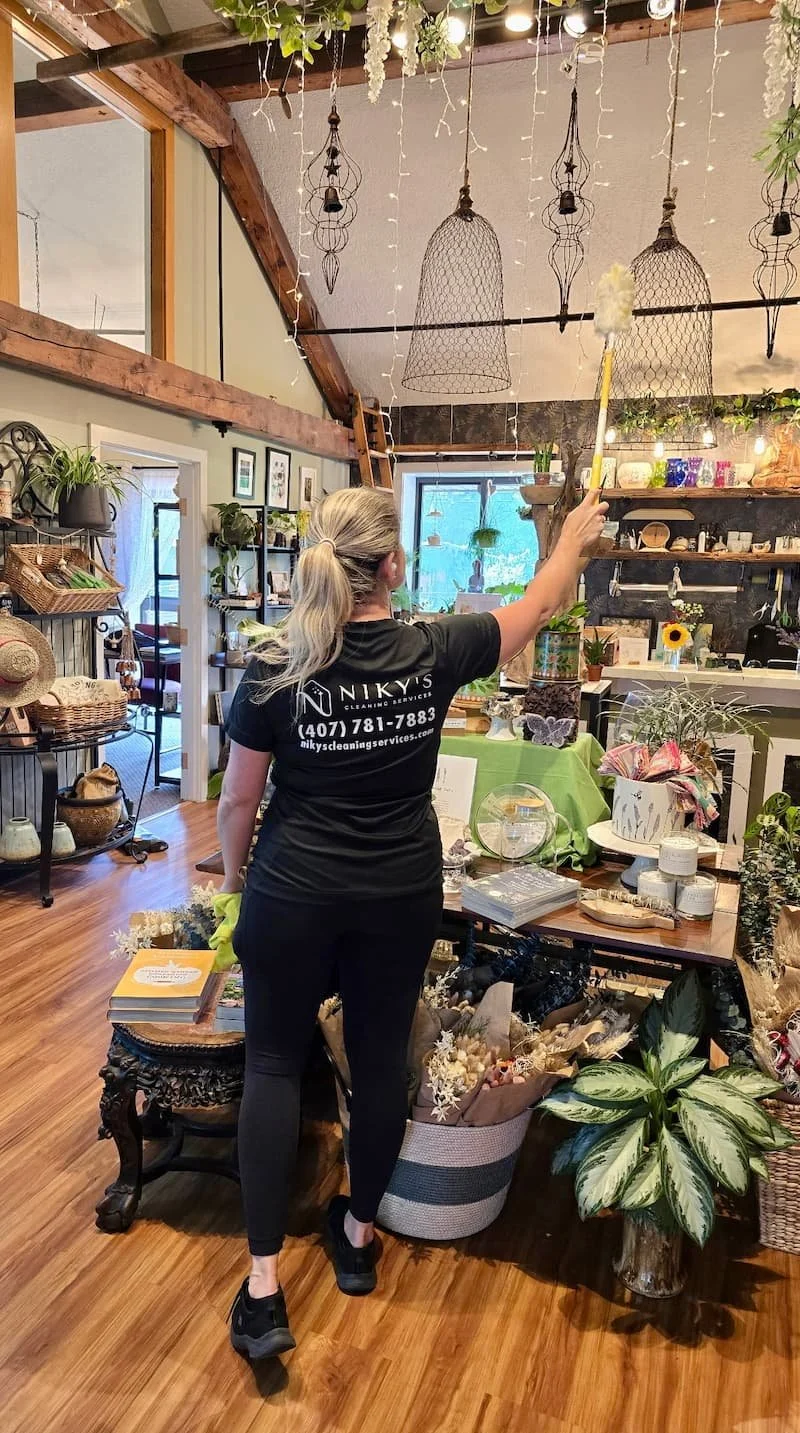 A woman cleaning a store interior with a duster. She has gray hair tied in a ponytail, wears black clothing with a logo on the back, and black sneakers. The store has wooden floors, decorated with plants, shelves with various items, and hanging light fixtures.