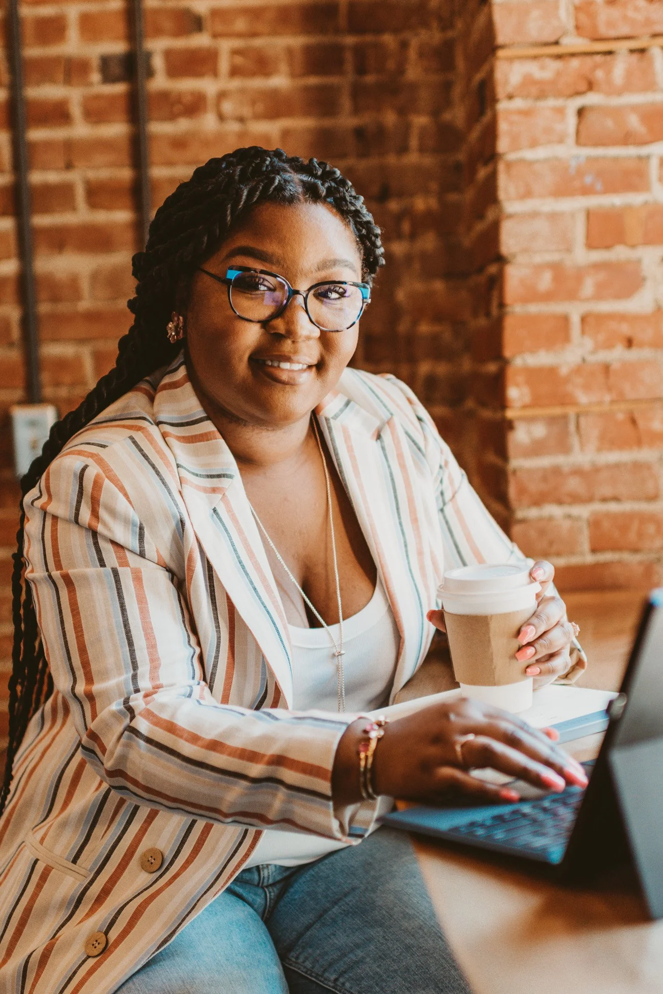 A woman with glasses and braided hair smiling at the camera, holding a coffee cup, sitting at a table with a laptop in front of her, in a brick-walled cafe or office.