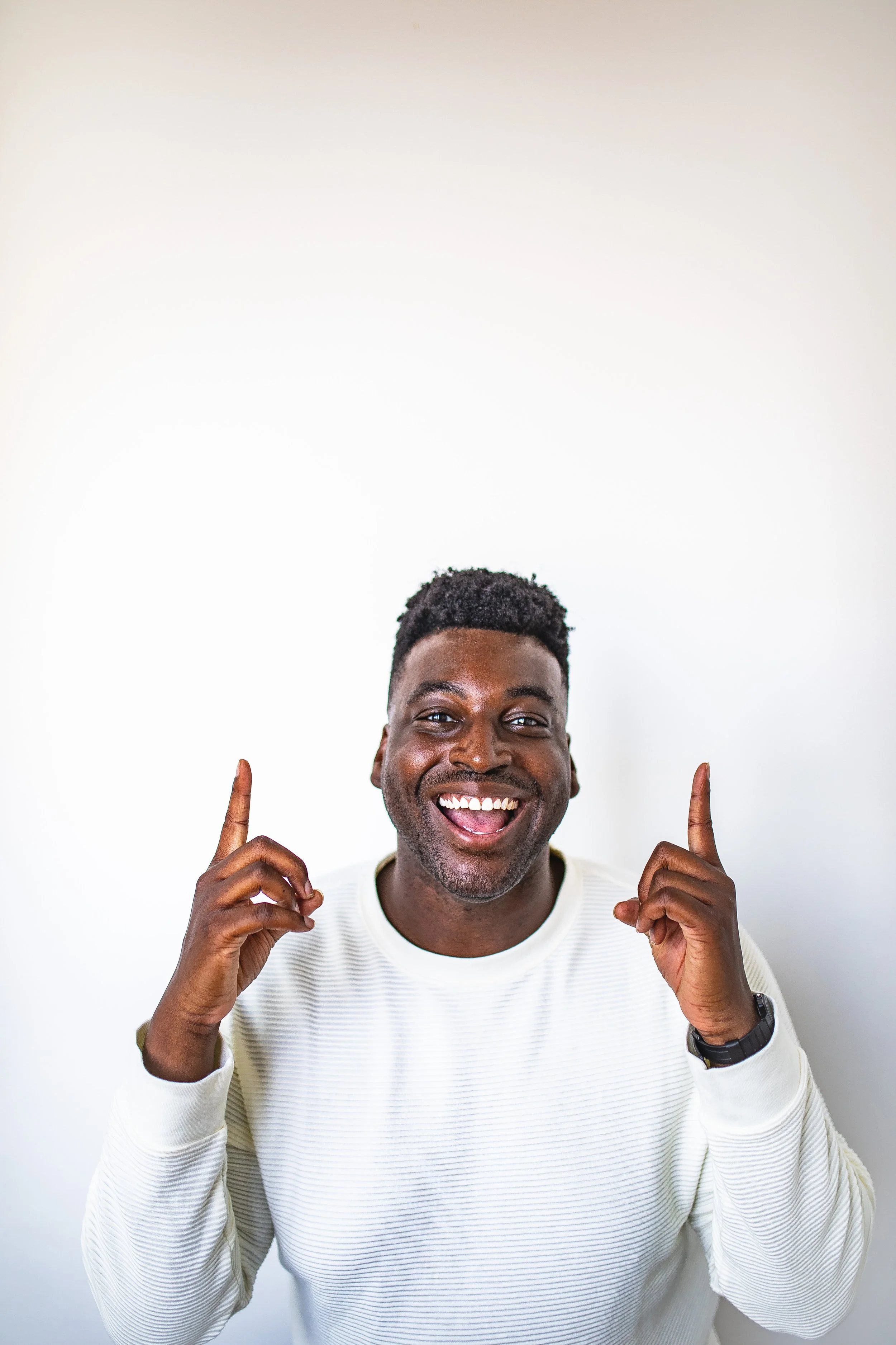 Smiling man with short curly hair pointing up with both hands, wearing a white long-sleeve shirt, against a plain white background.