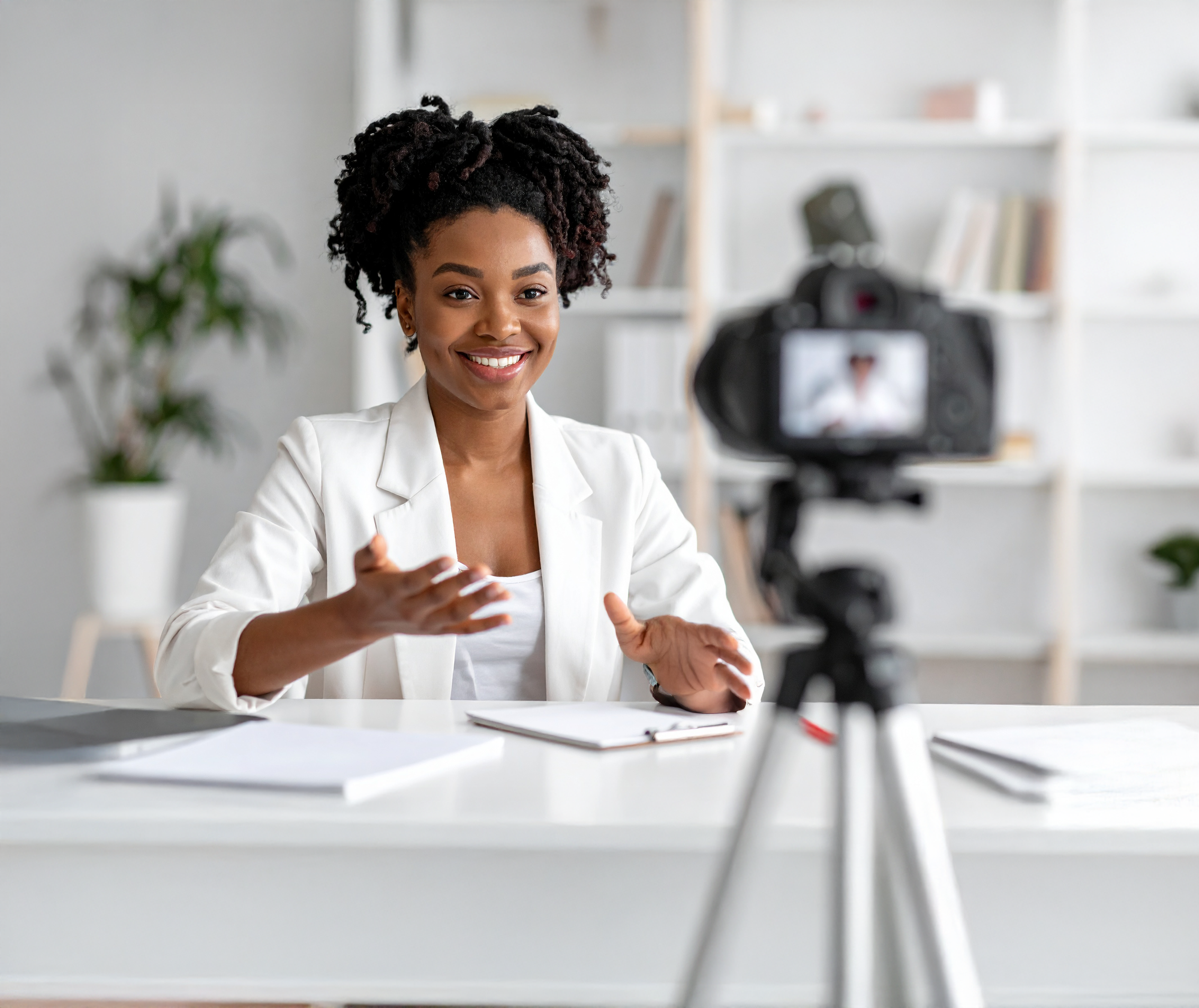 A woman with natural curly hair smiling and gesturing with her hands during a video recording in a bright, modern office setting.