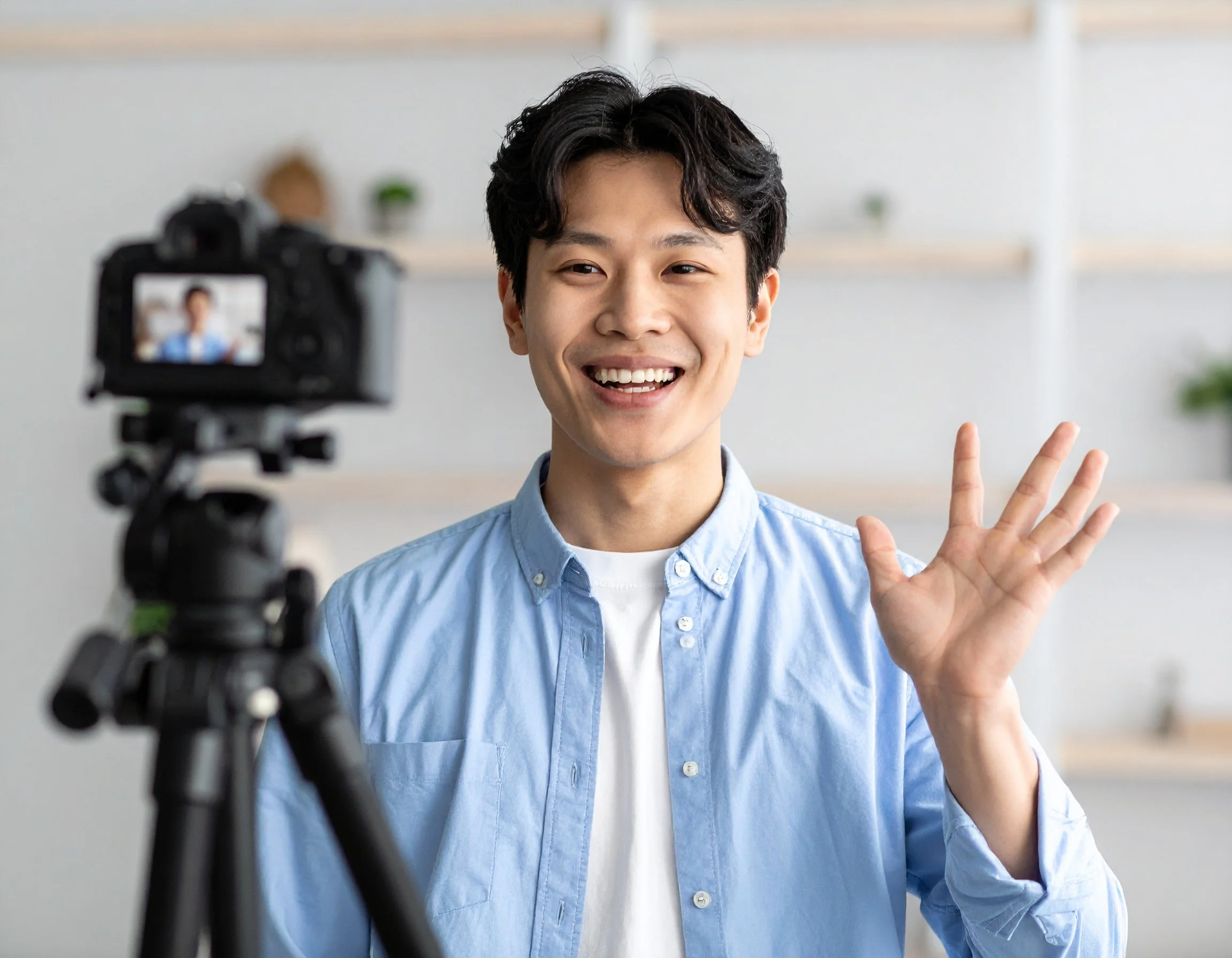 A young man is recording a video or live stream, smiling and waving at the camera, in a bright, modern indoor setting.