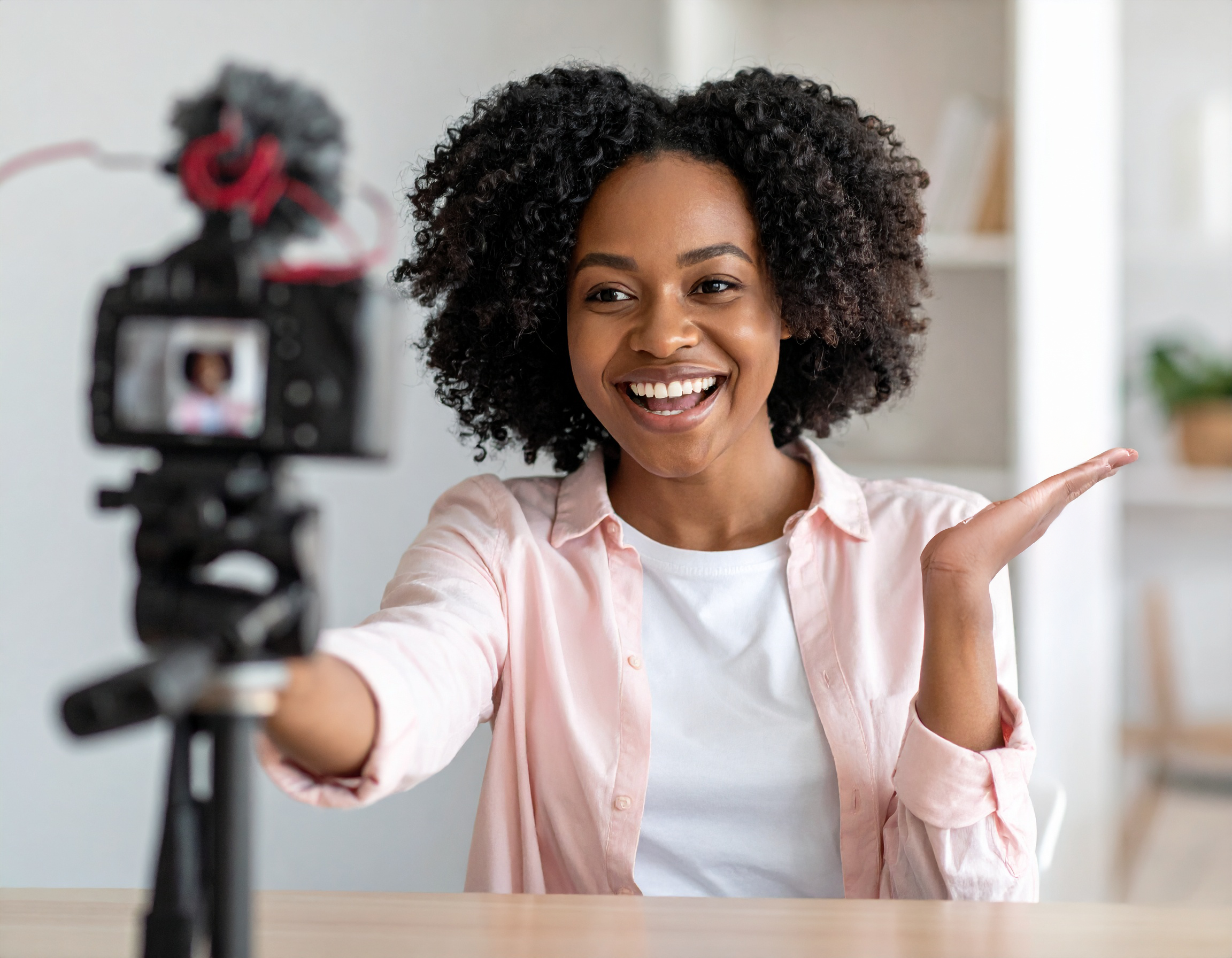 A woman with curly hair smiling and gesturing with her hand while recording a video in front of a camera.