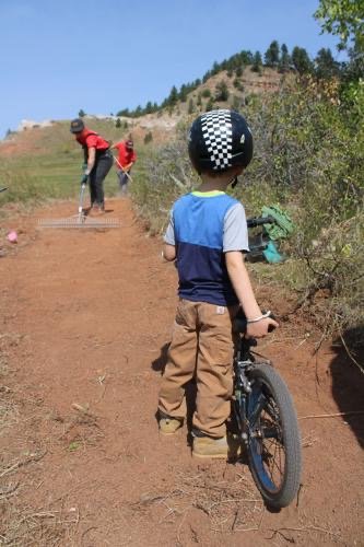 Child in a helmet and casual clothes riding a black bicycle on a dirt trail with two other children in the background hiking up the trail on a sunny day in a hilly outdoor area with trees.