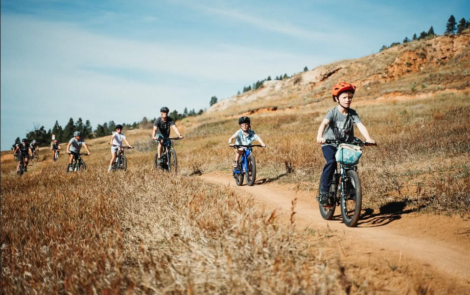 A group of children and teenagers riding mountain bikes along a dirt trail in a hilly, grassy landscape under a blue sky.