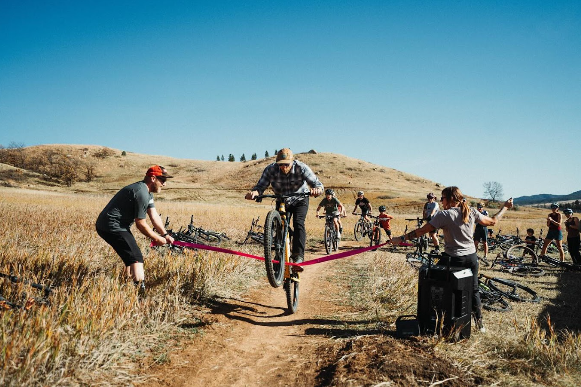 Person jump riding a mountain bike in a dirt field during a race or event, with spectators and other cyclists watching in the background.
