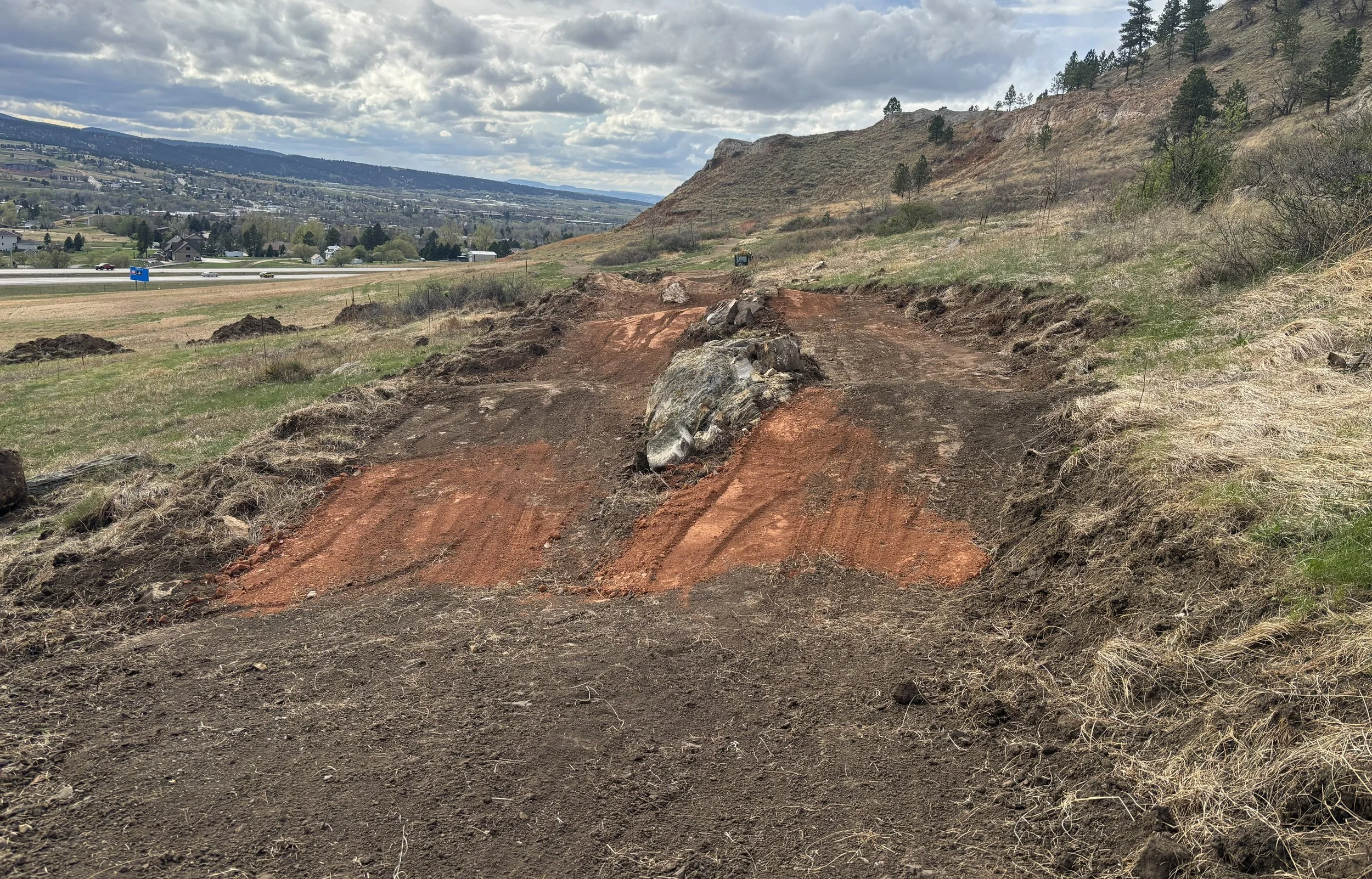 A dirt trail with rocks along the side, leading up a hillside with sparse vegetation, with an open view of a town below and mountains in the distance under a cloudy sky.
