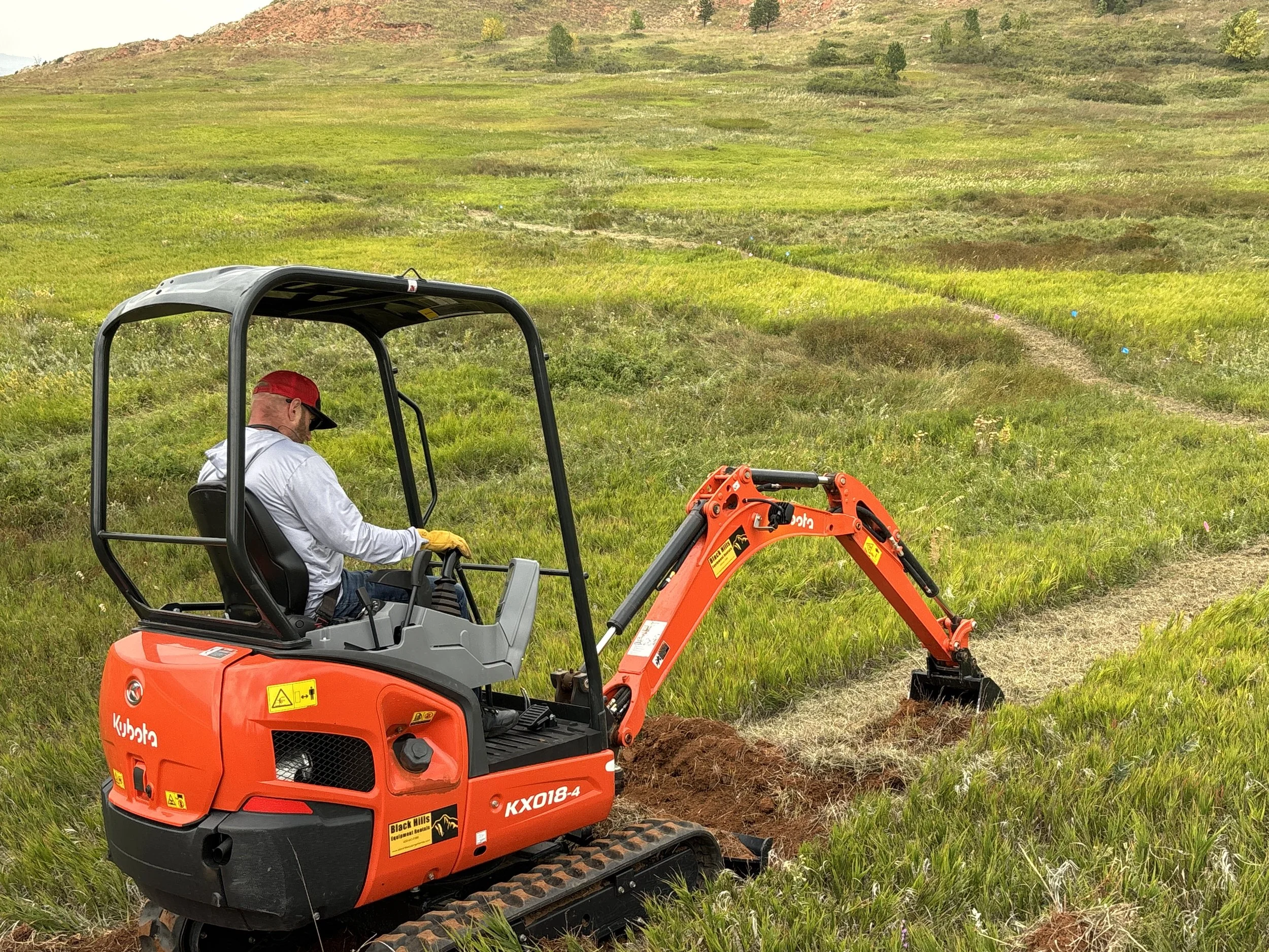 A man operating an orange Kubota compact excavator digging into the ground in a grassy field with rolling hills and small trees in the background.