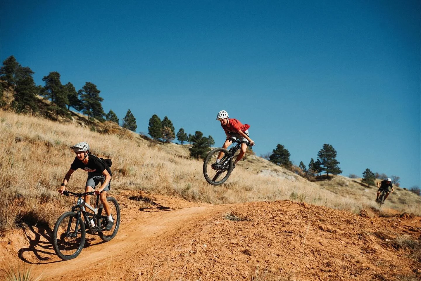 Three mountain bikers wearing helmets riding on a dirt trail through dry grassland with trees and blue sky in the background.