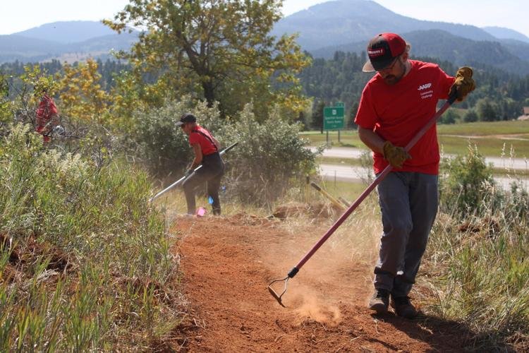 Two men working on a dirt trail, one in the foreground raking soil and the other in the background using a tool, in an outdoor area with trees, shrubs, and mountains in the distance.