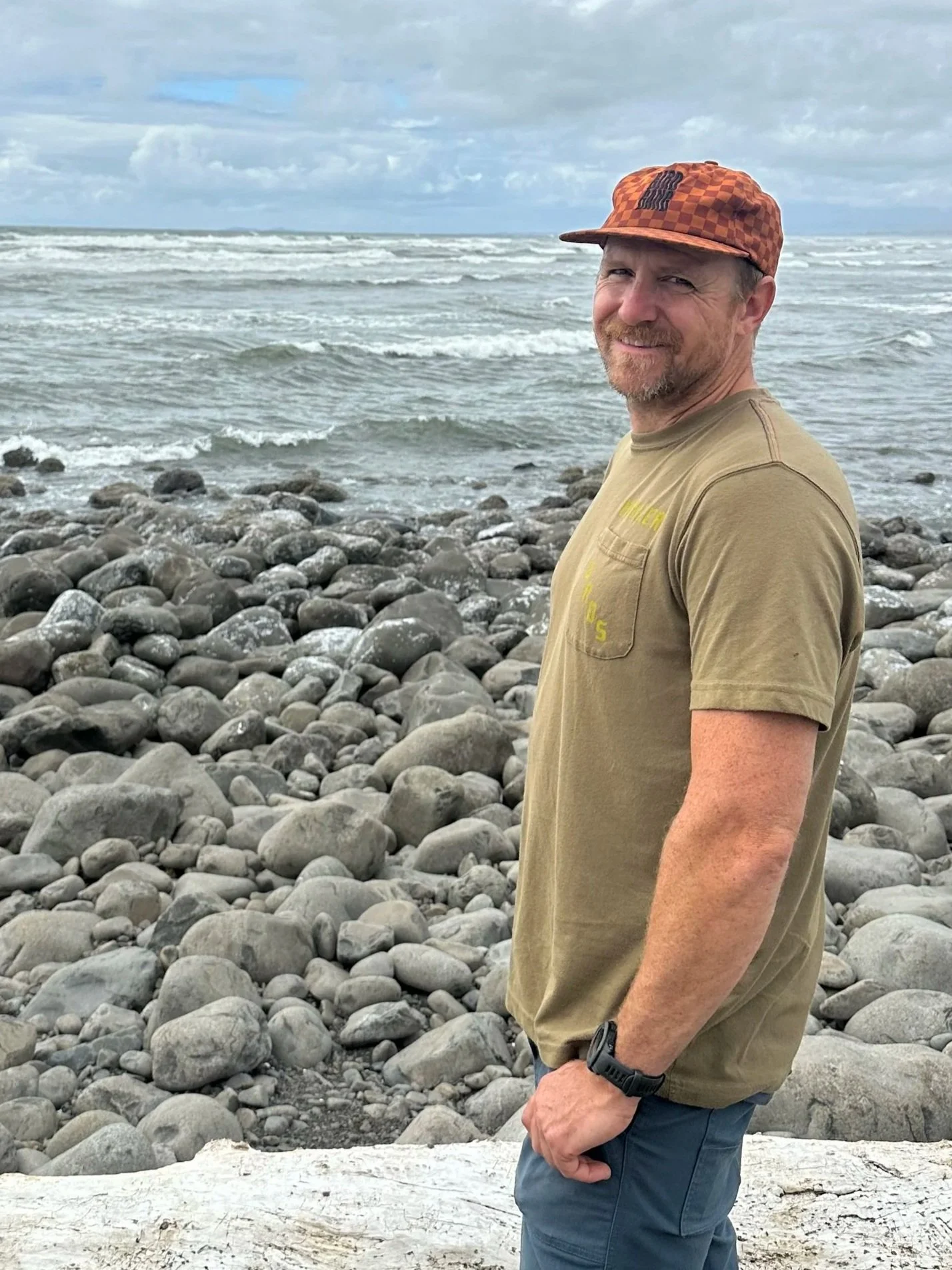 A man wearing a brown t-shirt, blue pants, a black watch, and an orange checkered cap stands on a rocky beach with the ocean and cloudy sky in the background.